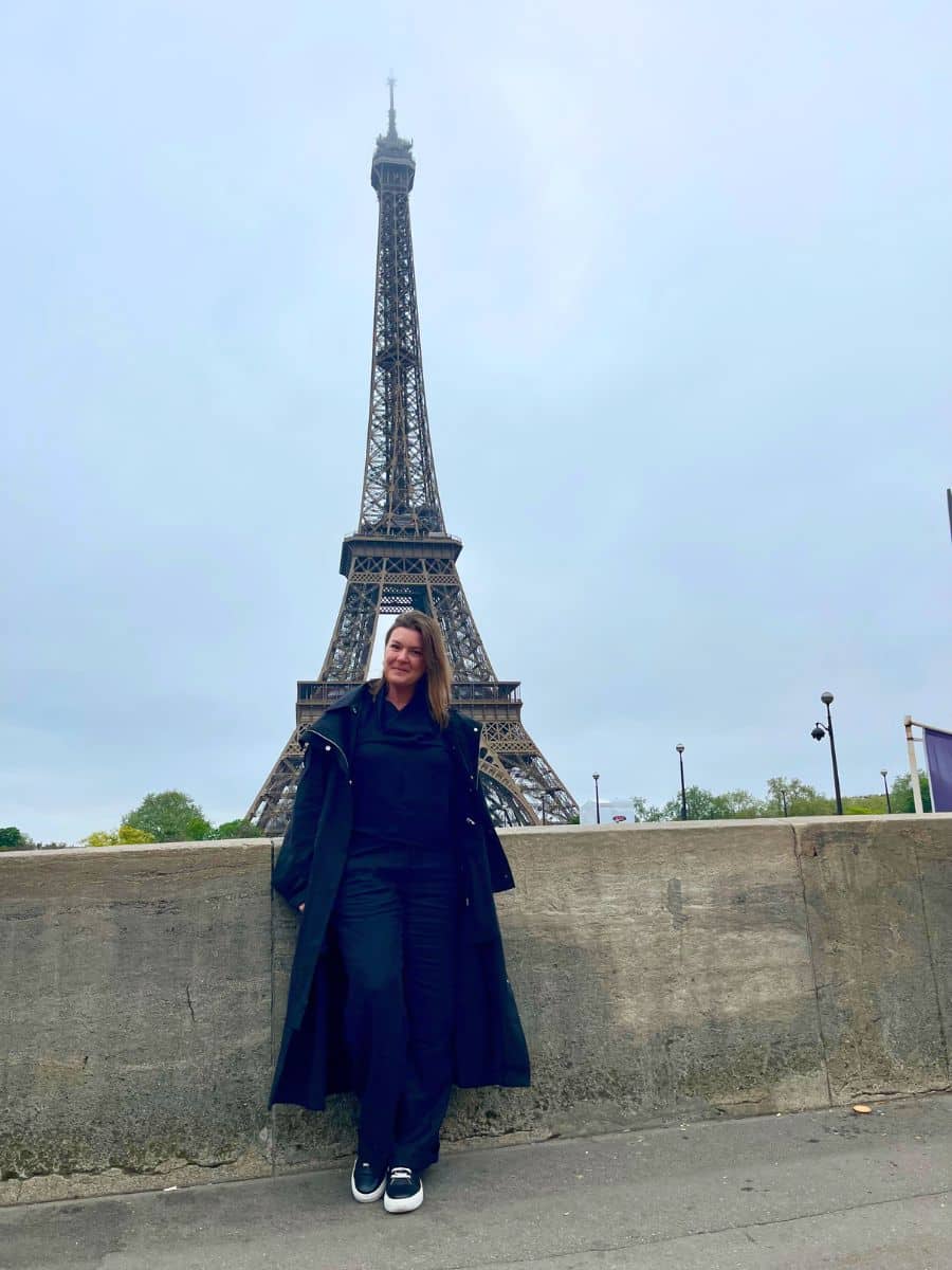 A solo female traveler, dressed in a stylish all-black outfit including a long coat, stands confidently in front of the Eiffel Tower in Paris. The overcast sky and empty surroundings add a serene quality to the scene, emphasizing the tower's majestic structure. She poses with a relaxed stance, smiling slightly, capturing a moment of her travel adventure in Paris.