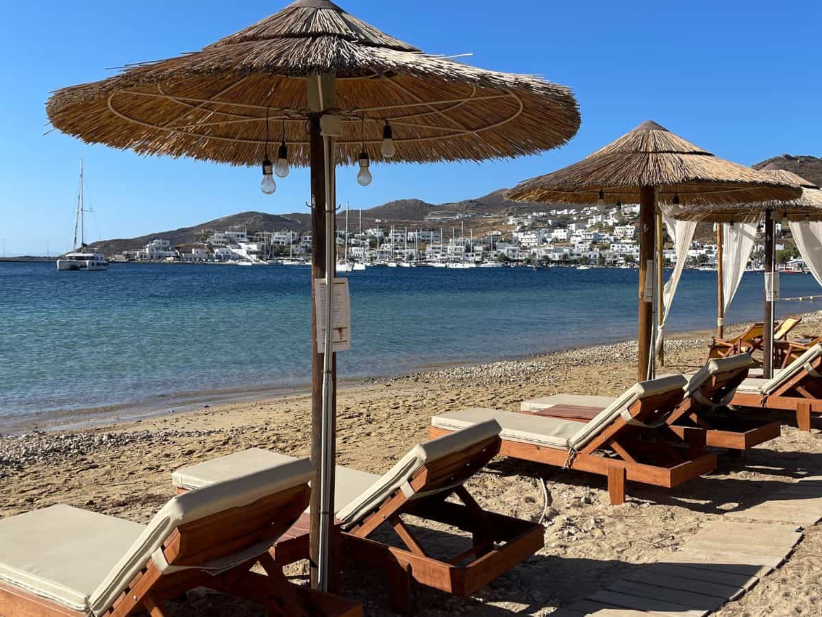 A tranquil beach scene with wooden sunbeds under thatched umbrellas on the sandy shore of Mykonos. The calm blue sea is in the background, with white buildings and boats moored in the harbor under a clear sky.