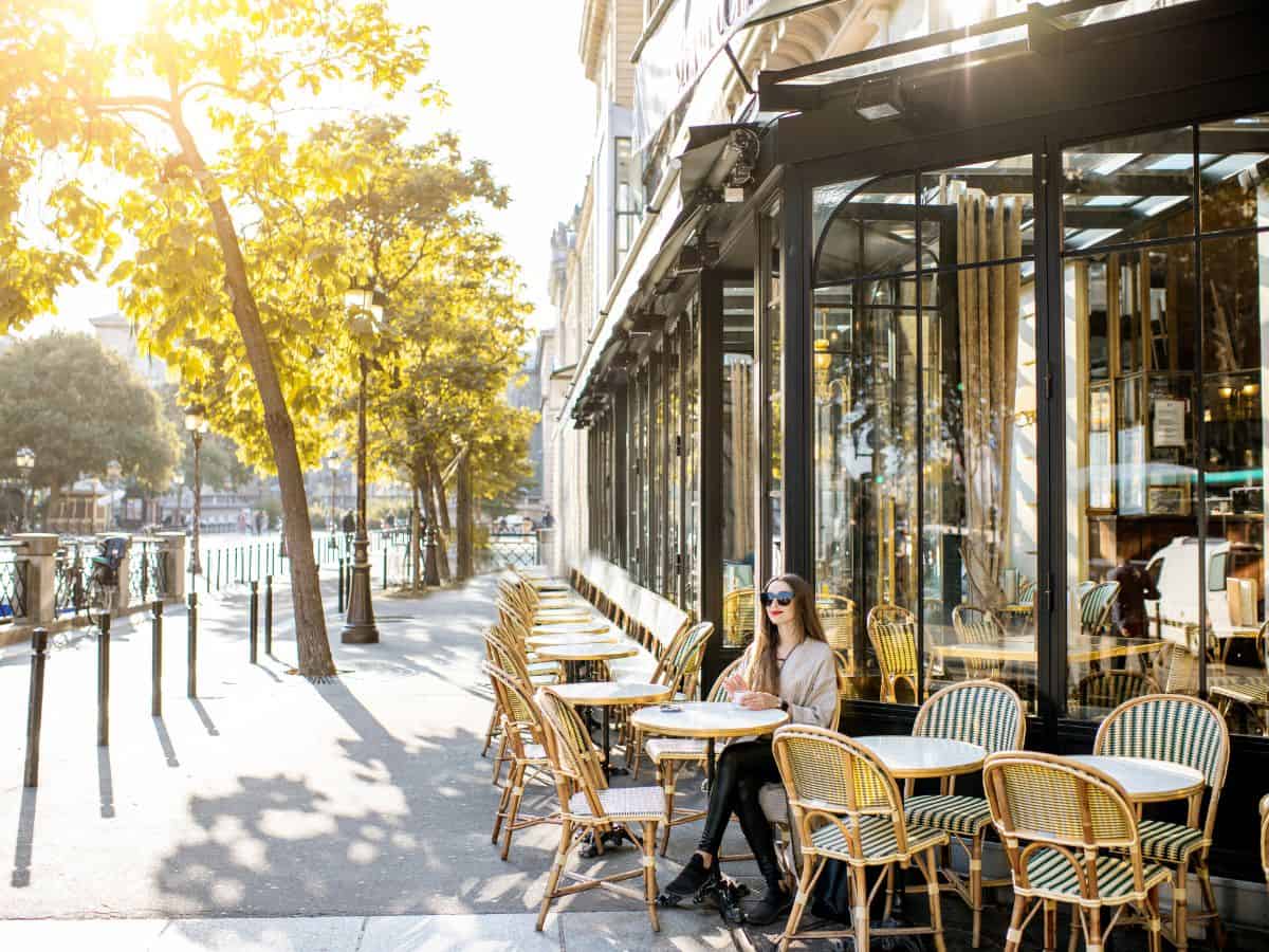 A woman enjoys a solo trip to Paris, seated at an outdoor cafe with rattan chairs, basking in the morning light. The street is quiet, lined with trees, evoking the relaxed pace of Parisian life.