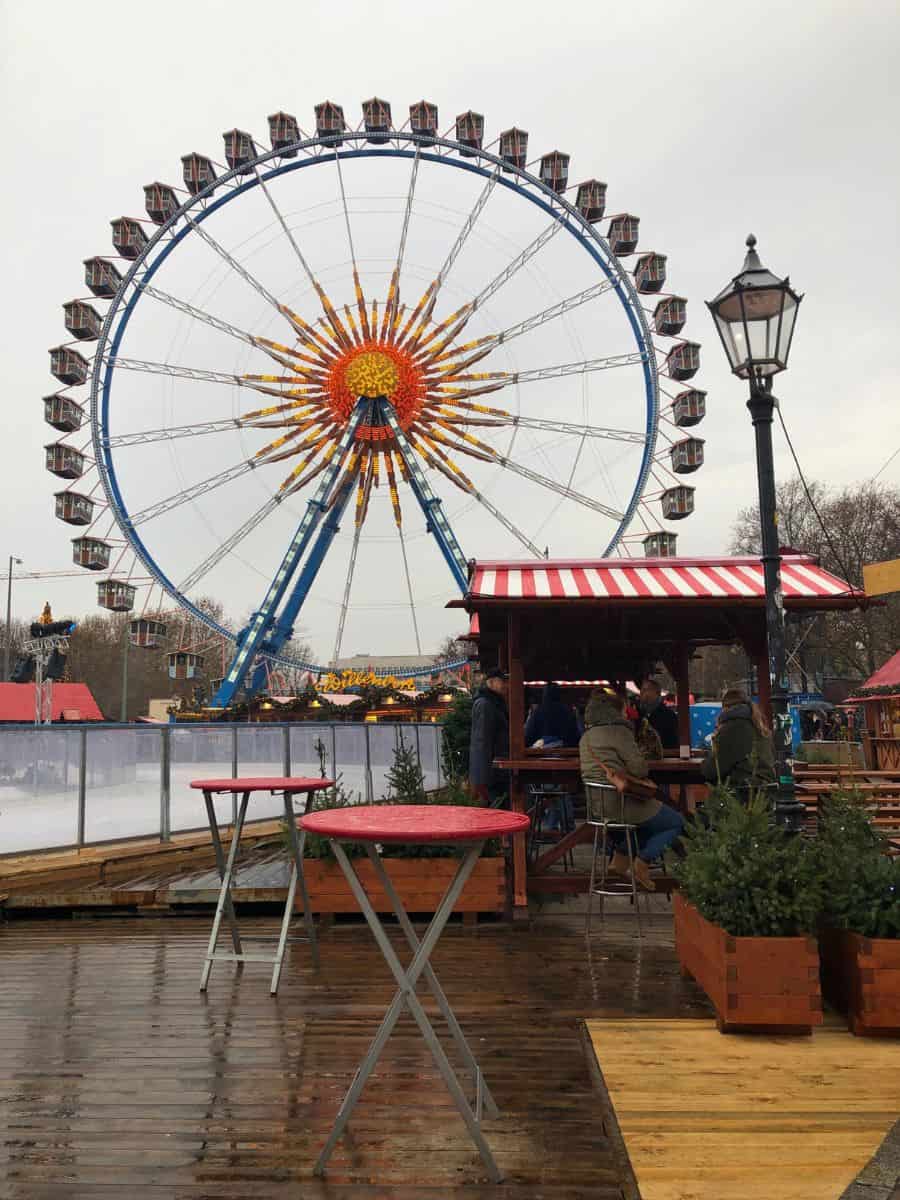 A giant Ferris wheel with vibrant blue supports and a sunburst-patterned center stands tall against the overcast sky at a Berlin Christmas Market, with visitors enjoying the festivities from nearby rustic wooden seating areas.