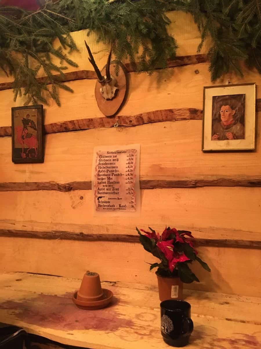 A cozy corner of a Berlin Christmas market stall, with rustic wooden walls adorned with a small antler trophy, vintage paintings, and a menu of traditional German holiday beverages, complemented by a poinsettia plant and a ceramic mug on a wooden bench.