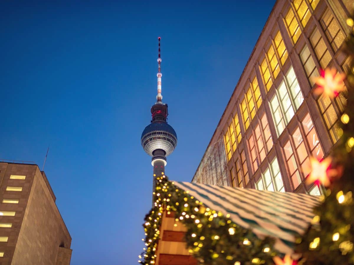 Twilight view of the iconic Berlin TV Tower rising majestically above a Christmas market, with the foreground showcasing a festively lit stall roof adorned with a garland and a glimpse of a warmly glowing building window.