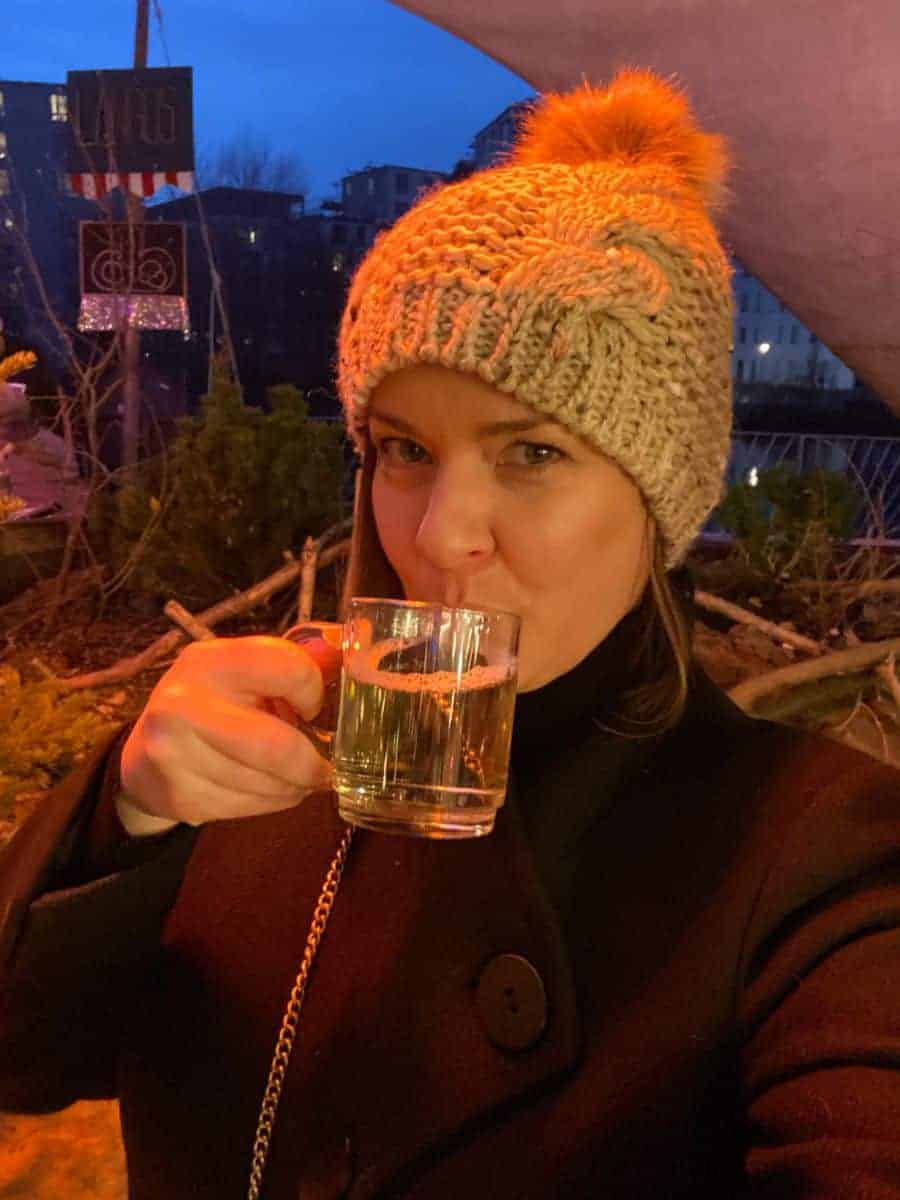 A woman in a warm knitted hat sips Glühwein at a Berlin Christmas market, her enjoyment evident in her expression, with the evening's ambient lighting softly illuminating the scene.