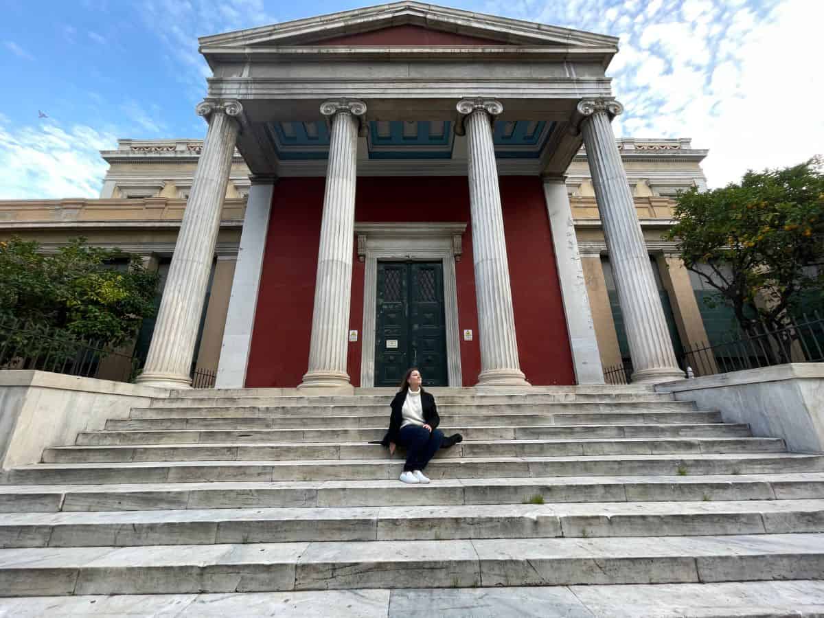 a woman living in athens sitting on the steps of the museum.