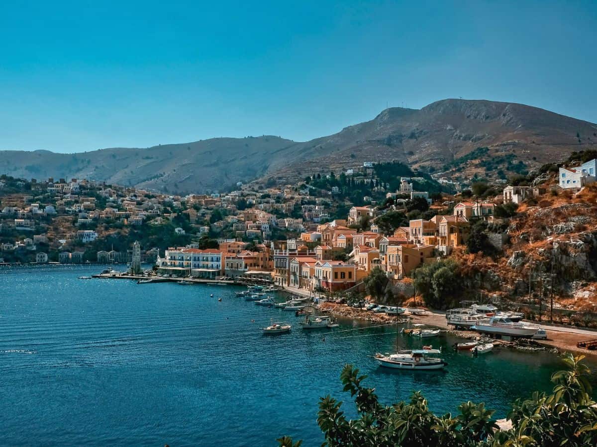 Panoramic view of a tranquil harbor on a Greek island, showcasing one of the hottest destinations to visit in May, with colorful buildings cascading down the hillside and boats gently bobbing in the calm blue sea.