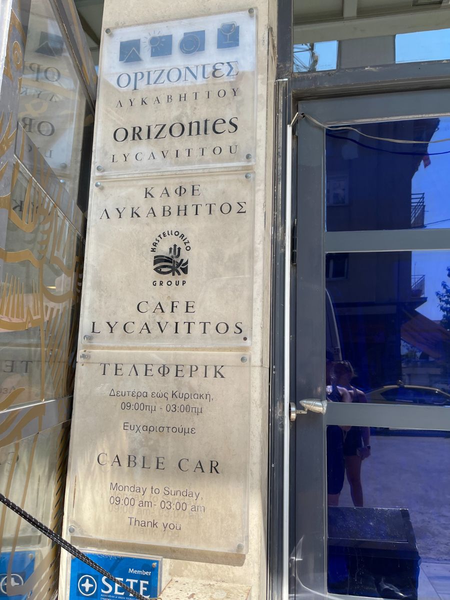 Sign at the entrance of the cable car to Mount Lycabettus in Athens. It lists "Orizontes Lycavittou" and "Cafe Lycavittos" along with operating hours for the cable car: Monday to Sunday, 9:00 am to 3:00 am. The text is in Greek, English, and Russian.