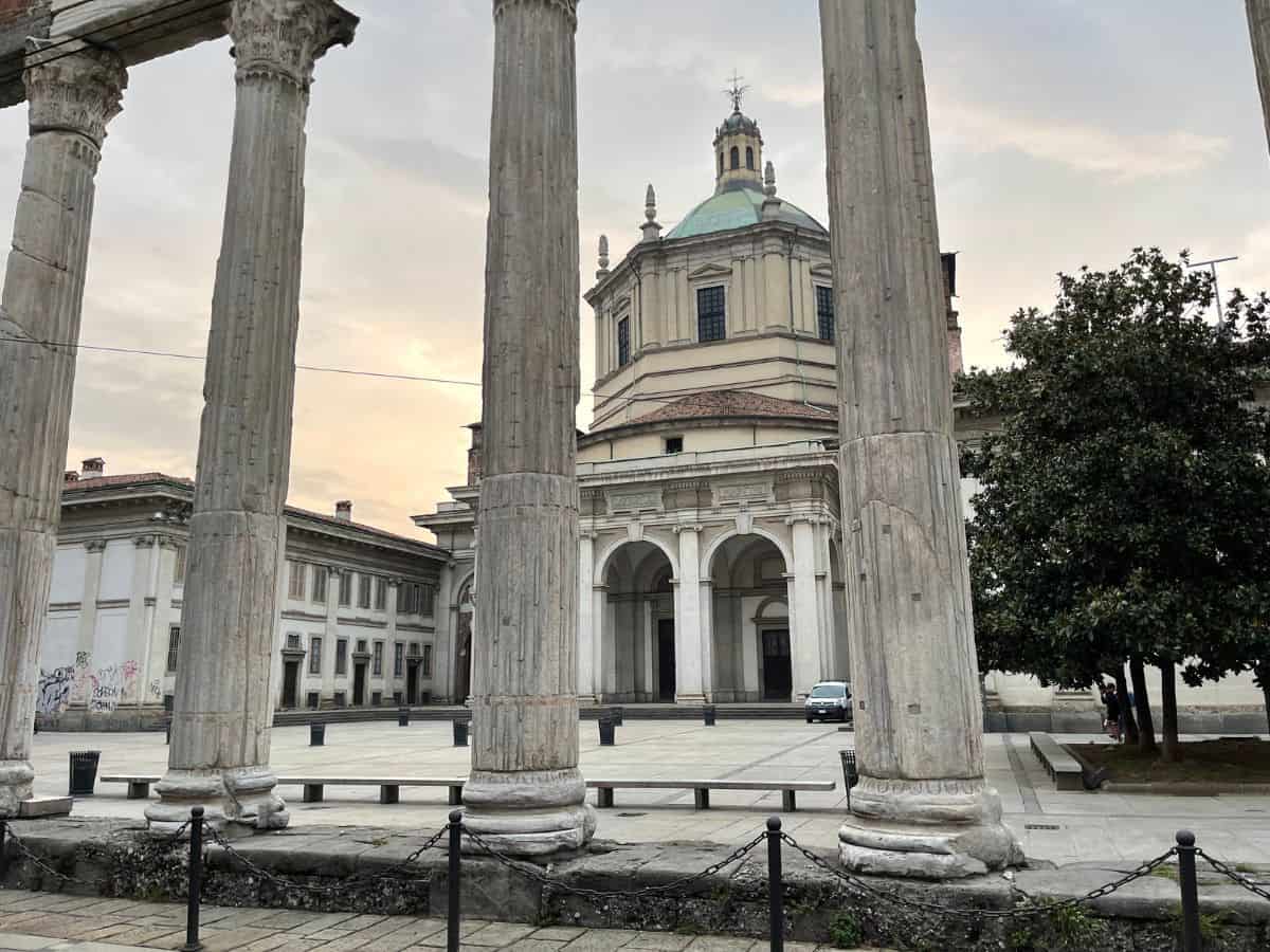 View of the Basilica di San Lorenzo in Milan framed by the ancient Columns of San Lorenzo. The classical architecture of the basilica, with its dome and facade, contrasts with the weathered Corinthian columns. An overcast sky looms above the quiet piazza in the early morning light.