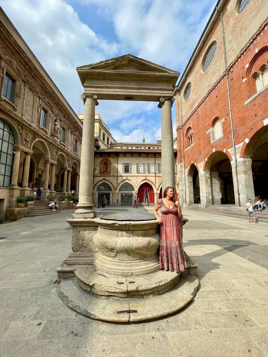 A woman in a patterned dress stands thoughtfully by an ancient stone well, topped with a classical canopy, in the picturesque courtyard of Milan's Statale University. The university's renaissance architecture, complete with arched walkways and historic frescoes, surrounds the tranquil square under a bright blue sky