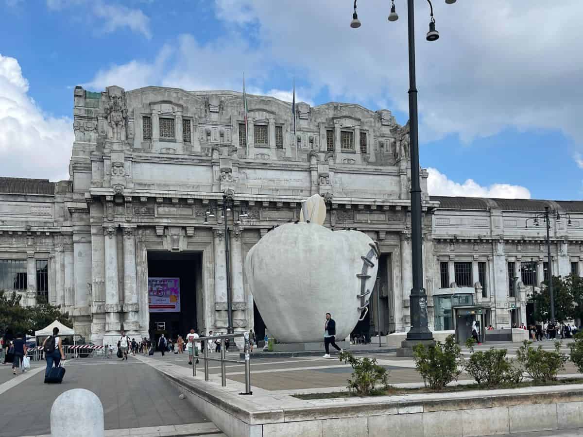 Milan's Central Station facade, a monumental example of 20th-century architecture, with pedestrians and travelers milling about. In the foreground, a large, striking sculpture of an apple with a zipper partially open stands as a modern contrast to the historic building, under a partly cloudy sky.