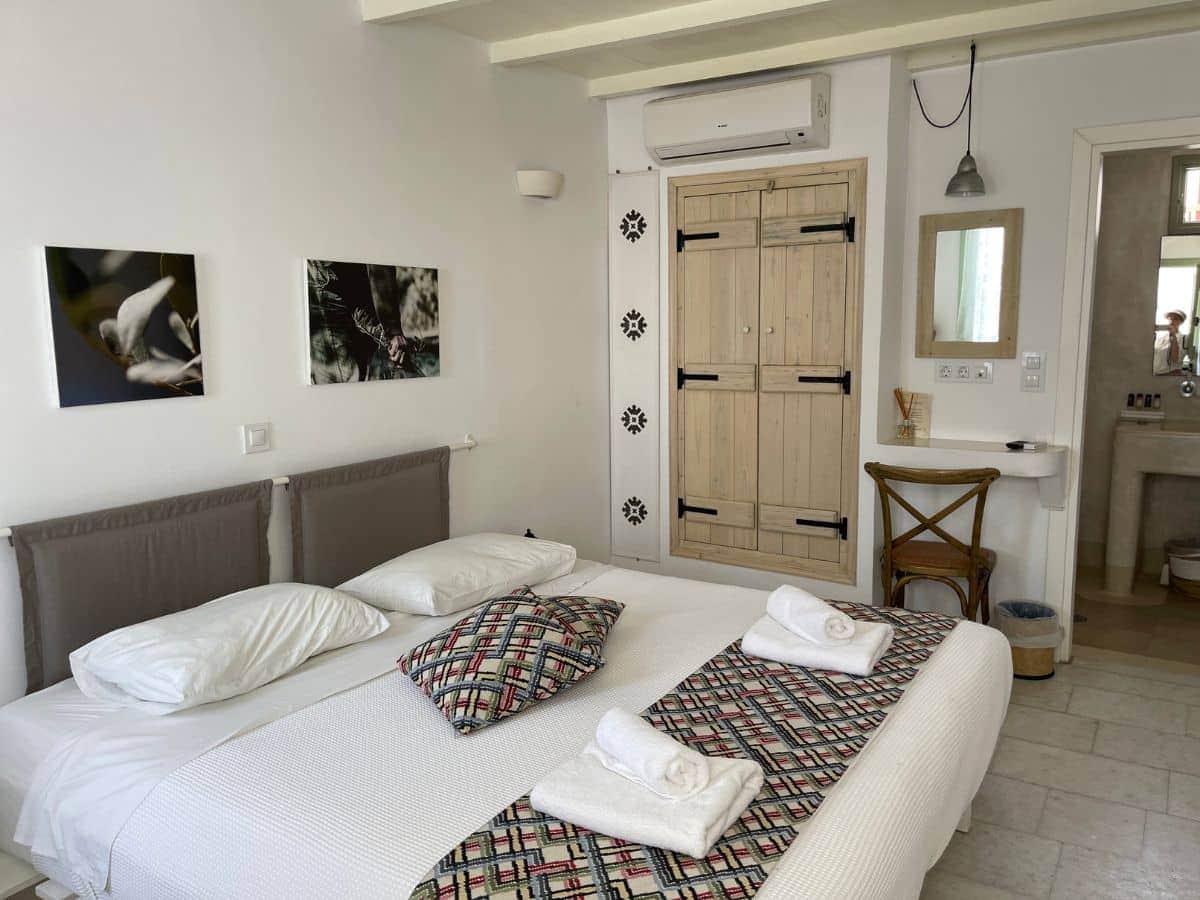 A modern and minimalist hotel room in Serifos or Naxos, featuring a large comfortable bed with crisp white linens and patterned throw pillows. The room is decorated with monochromatic botanical prints on the wall, a rustic wooden wardrobe, and a simple wooden chair beside a petite vanity area, which contributes to the room's clean and airy ambiance. A small pendant light hangs above, adding a touch of industrial charm to the cozy space.