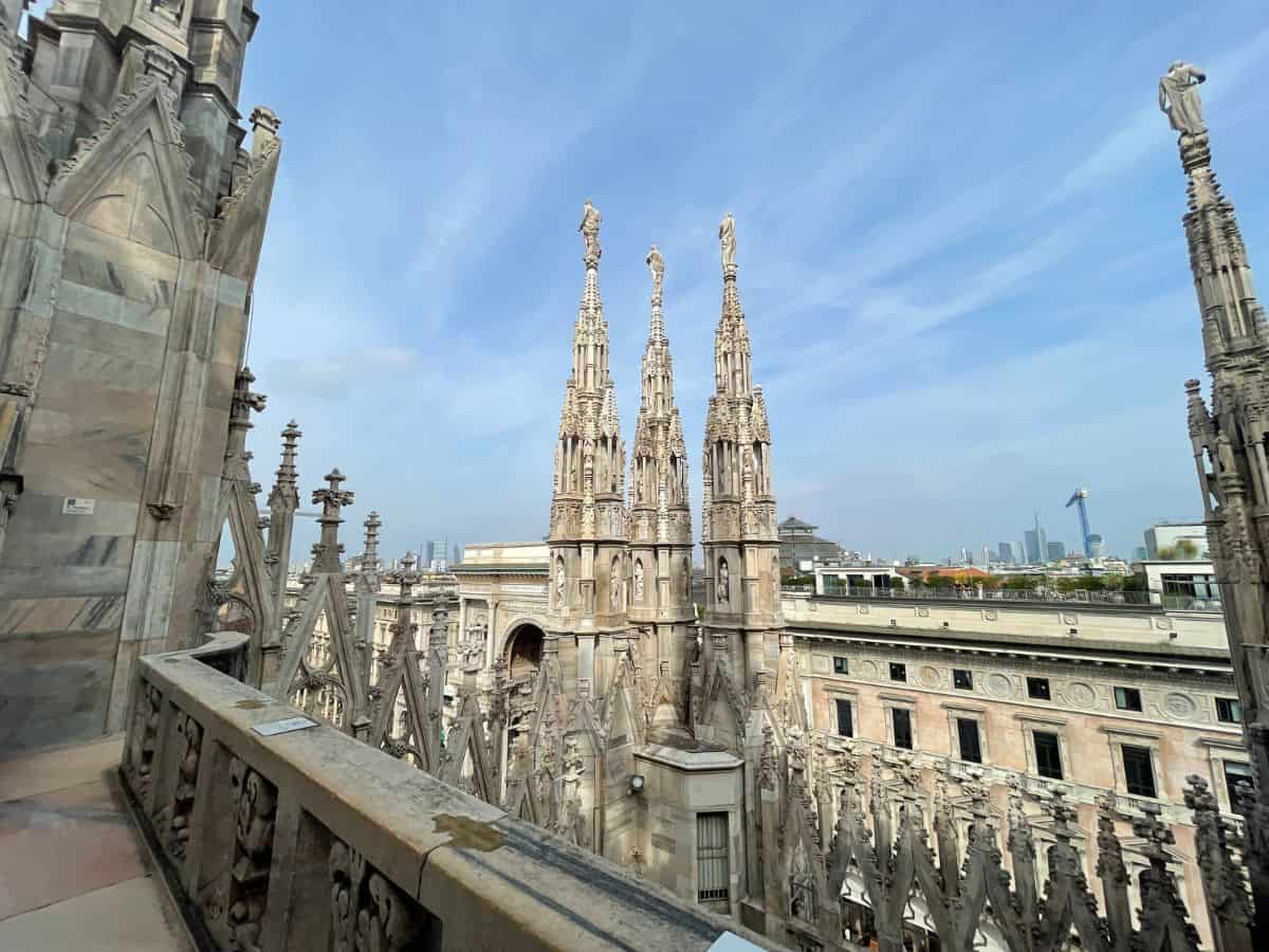 The intricate spires of the Milan Cathedral (Duomo di Milano) soar into the sky, showcasing the elaborate Gothic architecture. The foreground features the marble statues and ornate flying buttresses of the cathedral, with the cityscape stretching into the distance under a soft blue sky with wispy clouds.