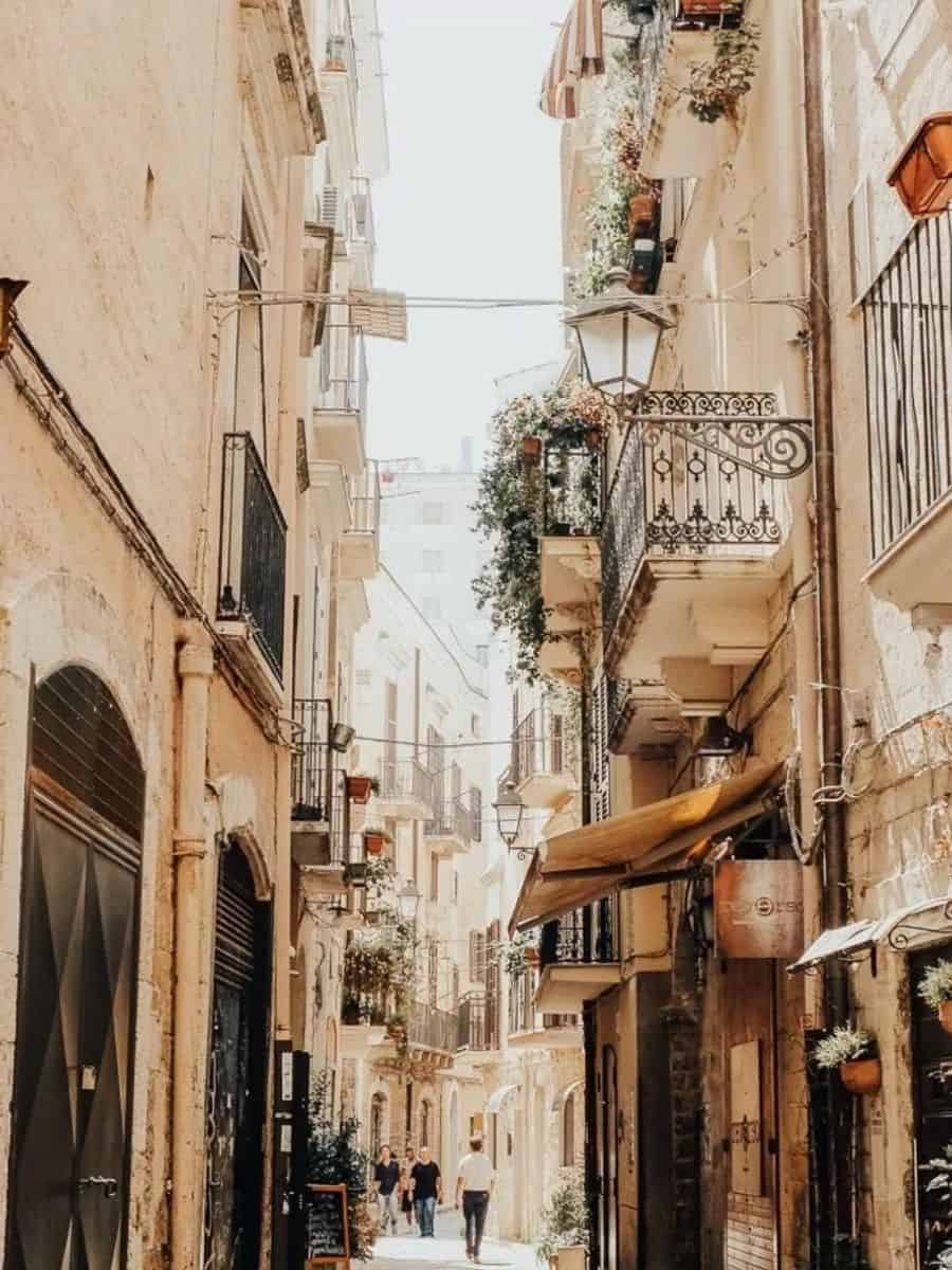 A narrow street in Bari, Italy, lined with old buildings and balconies adorned with plants. The street is filled with soft sunlight, giving it a warm, inviting appearance. People are seen walking in the distance.
