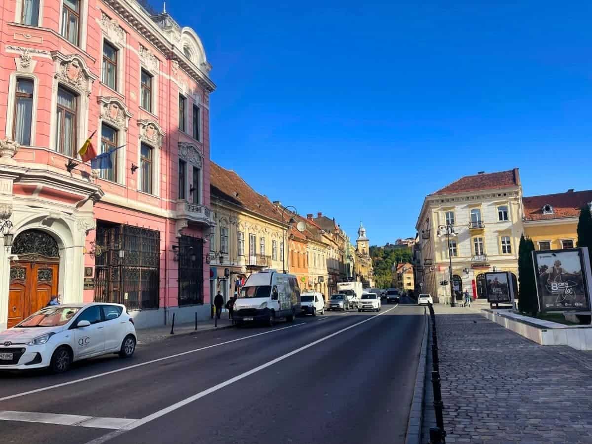 Street view in a Romanian city with a mix of classic and modern vehicles driving and parked along a cobblestone street. Historical buildings with European architecture line the road, displaying intricate façades and balconies, under a clear blue sky.