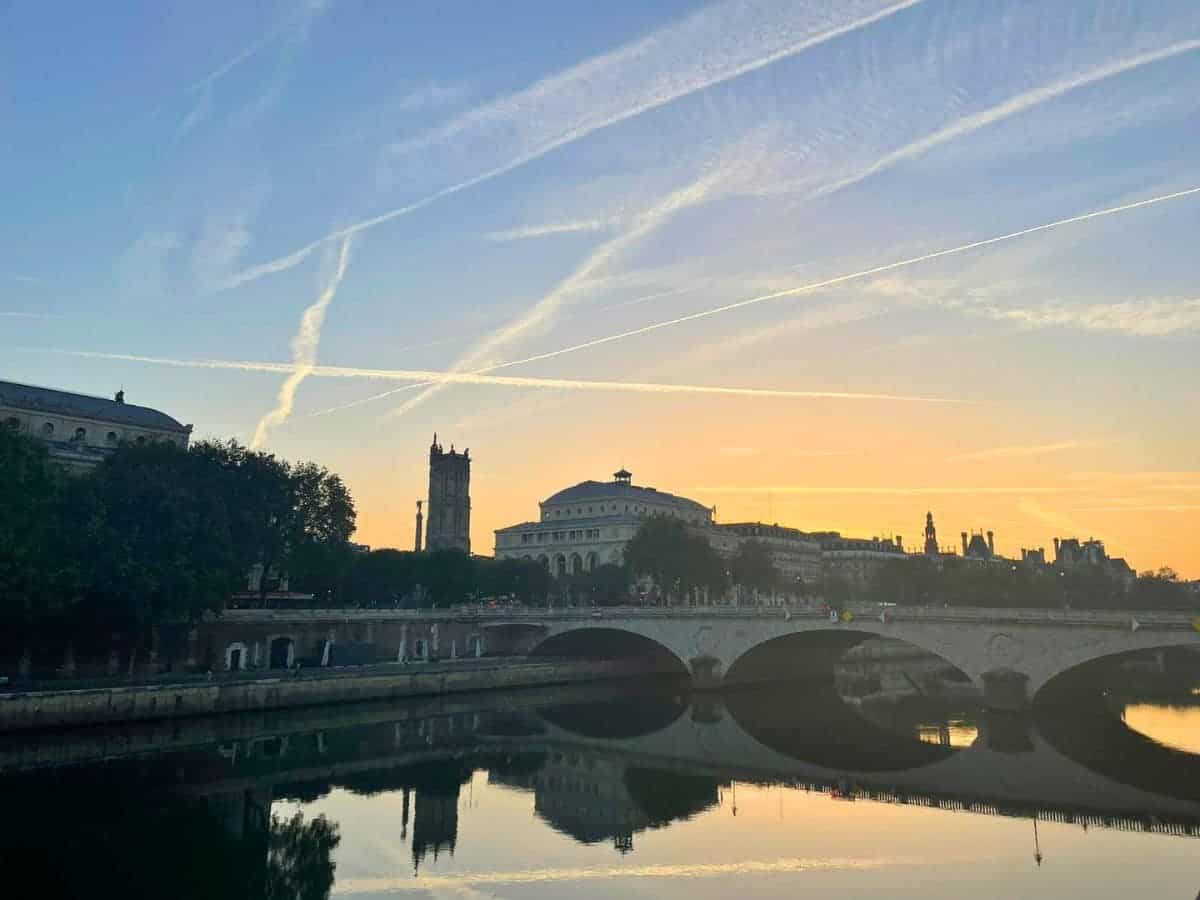 A tranquil scene captures the essence of a solo trip to Paris, featuring the silhouette of a historic riverside building and bridge against a sunrise sky with airplane trails crisscrossing above.