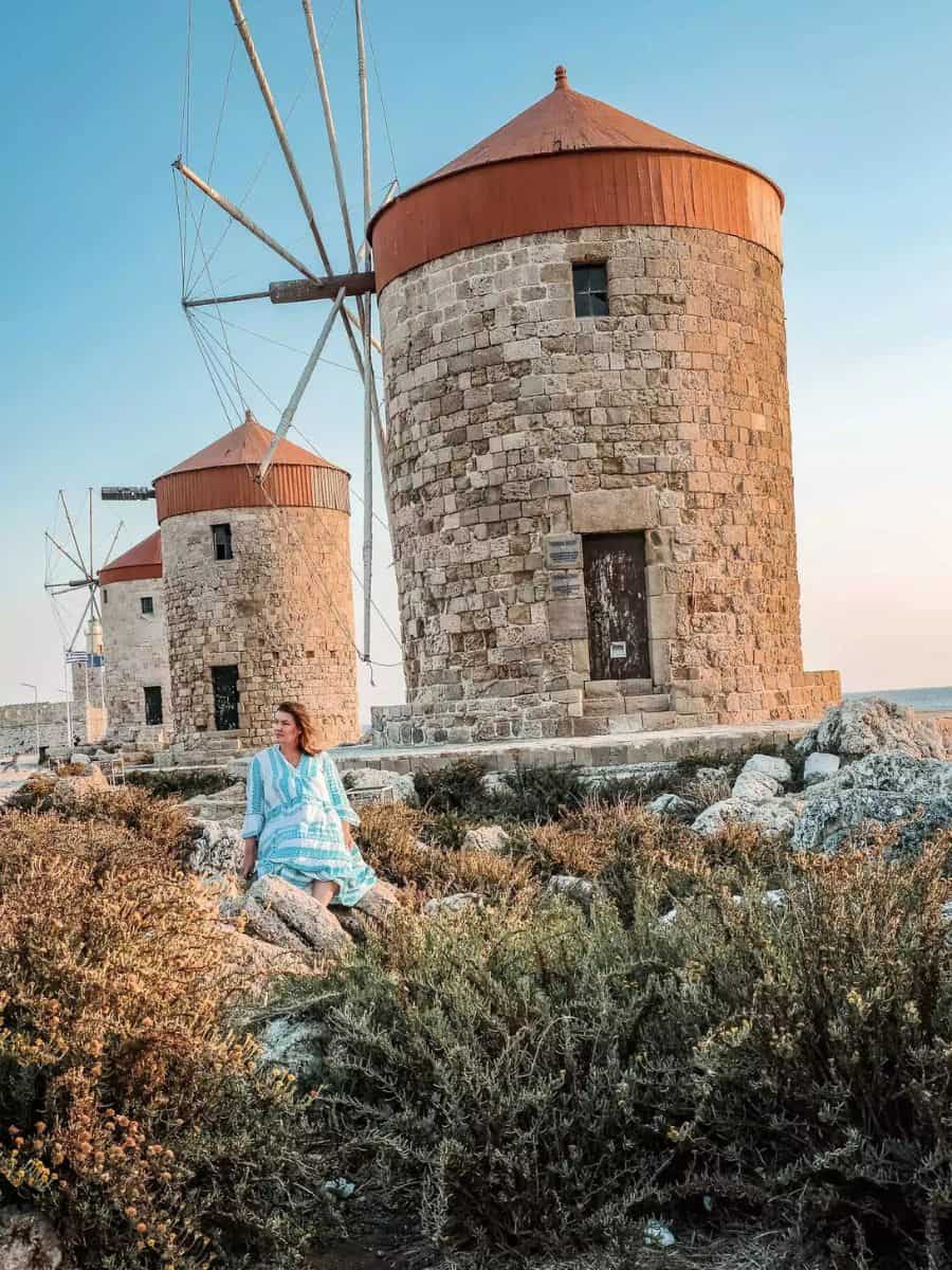 A woman  in a turquoise dress sits amidst wild herbs, with ancient stone windmills in the background on the Greek island of Rhodes, one of the hottest destinations for travel enthusiasts