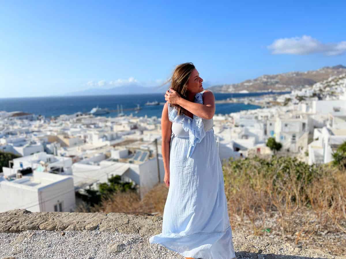 A woman in a flowing white dress stands atop a rocky outcrop overlooking the panoramic view of Mykonos town. The white buildings with blue accents and the calm sea extend into the horizon under a bright blue sky, evoking the quintessential beauty of the Greek islands.