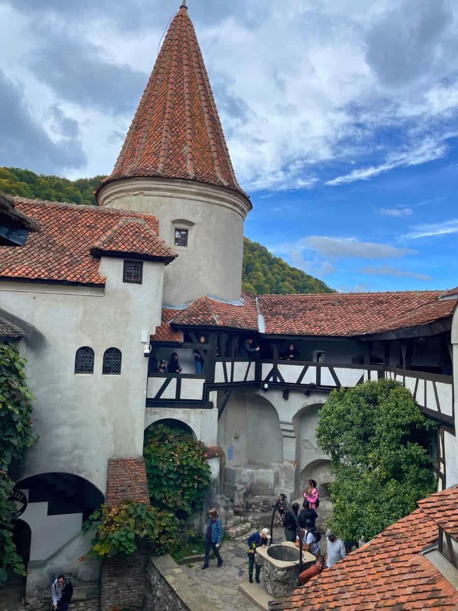Visitors explore the courtyard of Bran Castle, featuring a well, a stone tower with a conical roof, and the castle's white walls with timber framing, capturing a lively moment of cultural immersion.