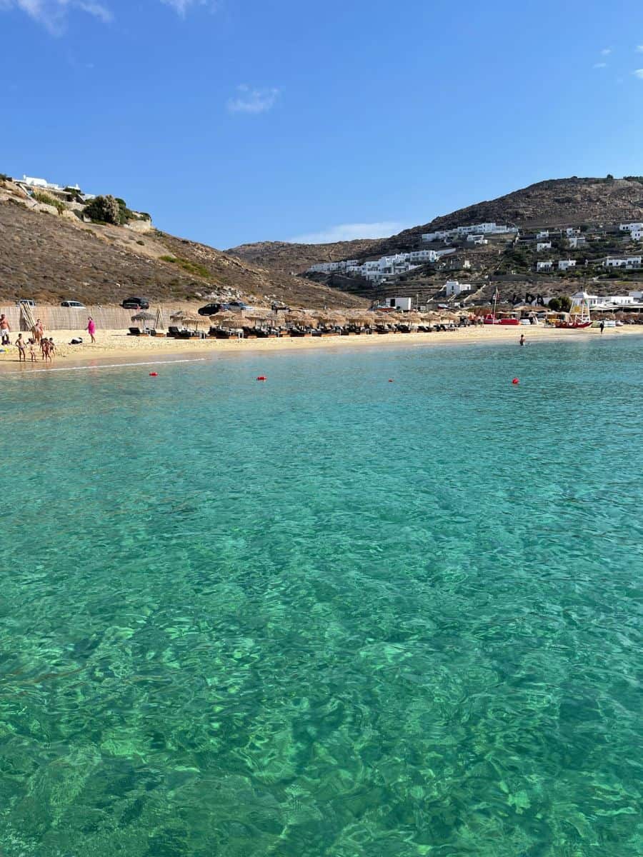 Crystal-clear turquoise waters edge onto a sandy beach in Mykonos, with sunbathers and hillside white buildings in the distance, showcasing the island's allure in May.