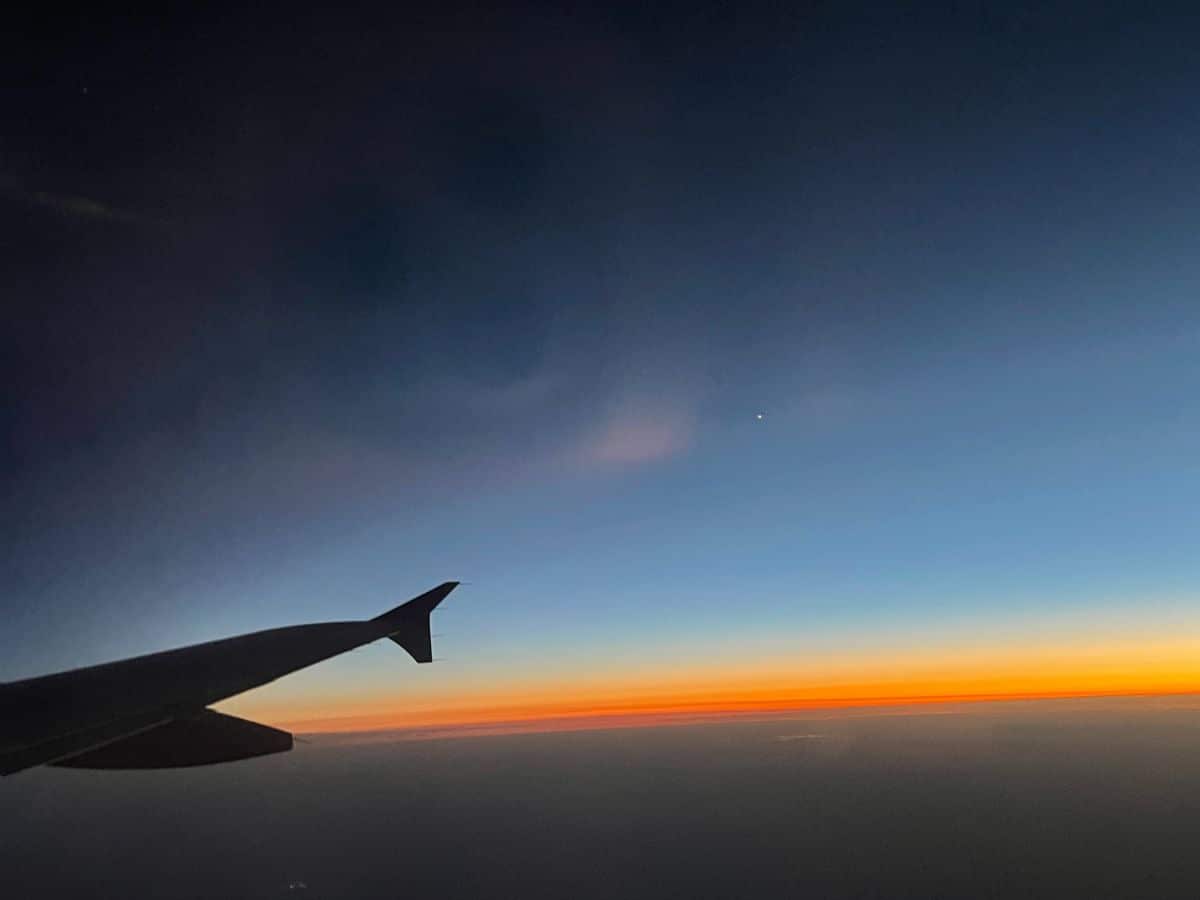 View from an airplane window during flight, capturing the wing silhouette against a breathtaking gradient of sunset colors blending into a darkening blue sky, evoking the peaceful solitude of travel at dusk.