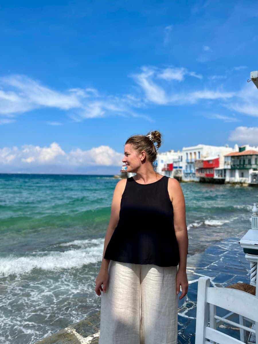 A woman in chic summer attire stands by the seafront in Mykonos, with the iconic whitewashed buildings in the background, under the bright Aegean sun, capturing the essence of a stylish getaway in May.