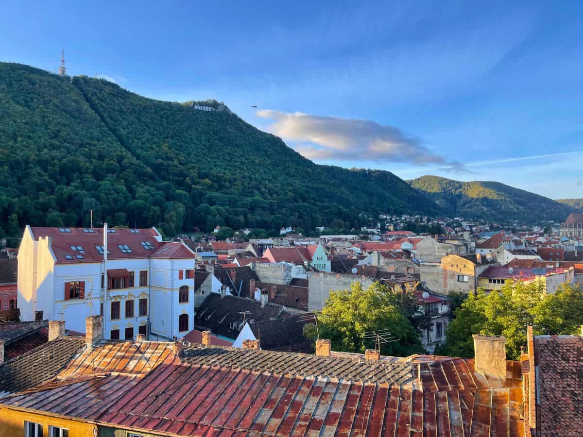 An elevated view over the red-tiled rooftops of Brasov, Romania, with the name 'BRASOV' spelled out on the lush, forested hillside in the background, conveying the picturesque charm of the town.