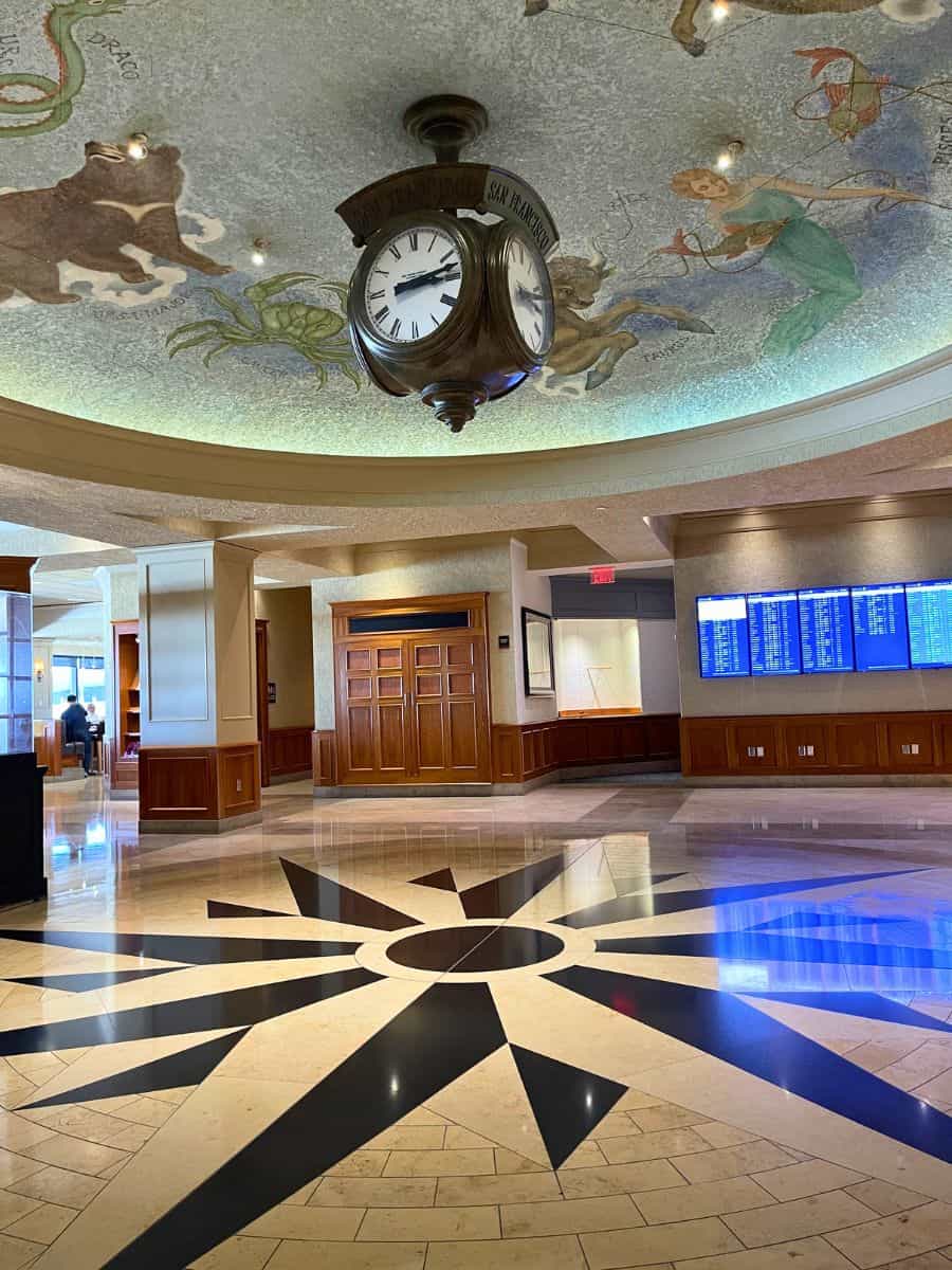 "Elegant airport lounge interior with a classic hanging clock showing the inscription 'San Francisco' and a painted ceiling depicting whimsical zodiac signs, while a flight information display board is visible in the background, inviting travelers to ponder if having a Priority Pass is worth the luxury and convenience offered.