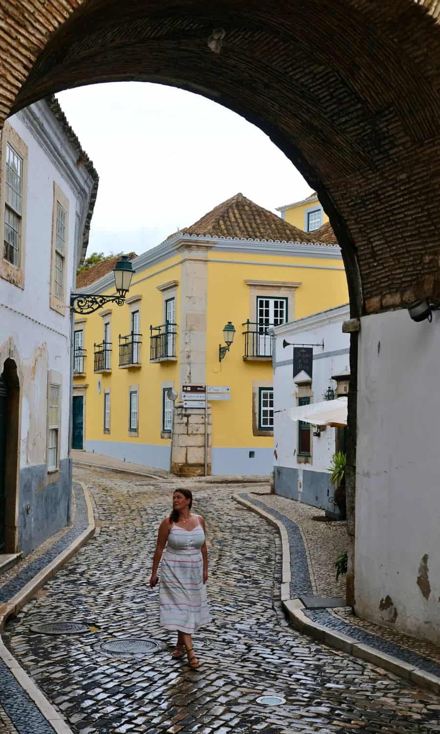 Solo female traveler walking under an archway on the cobblestone streets of Faro, surrounded by classic Portuguese architecture with a hint of vibrant yellow on the building facades.