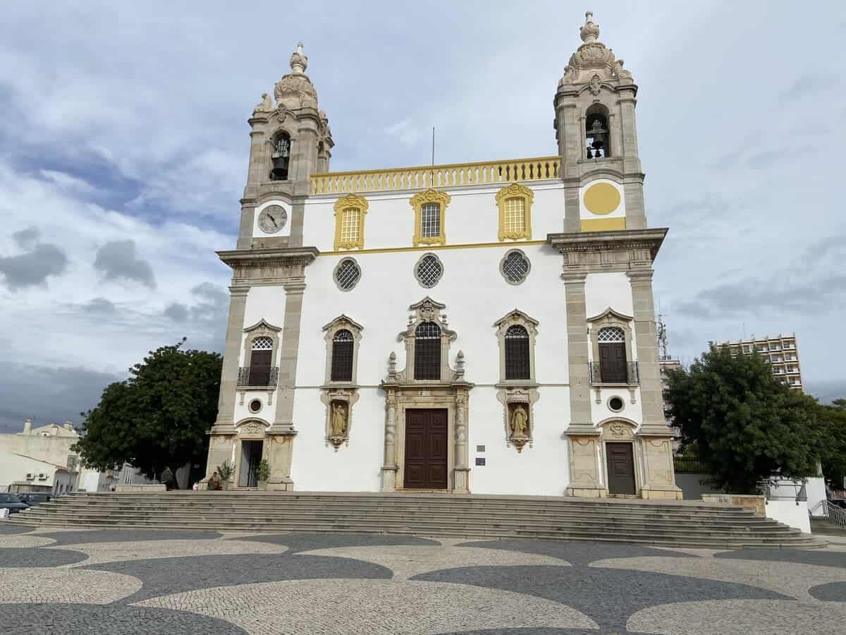 A striking white church with ornate baroque features and yellow trim stands under a cloudy sky in Faro, inviting contemplation and admiration