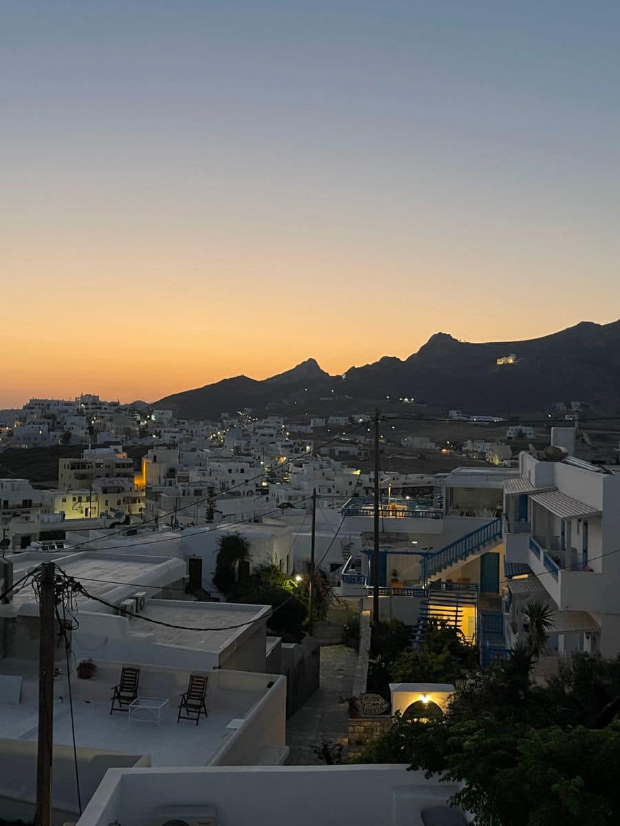 Twilight descends on Naxos, with a golden sunset backdrop silhouetting the mountains and casting a warm glow over the white cubic buildings, offering a tranquil evening scene.