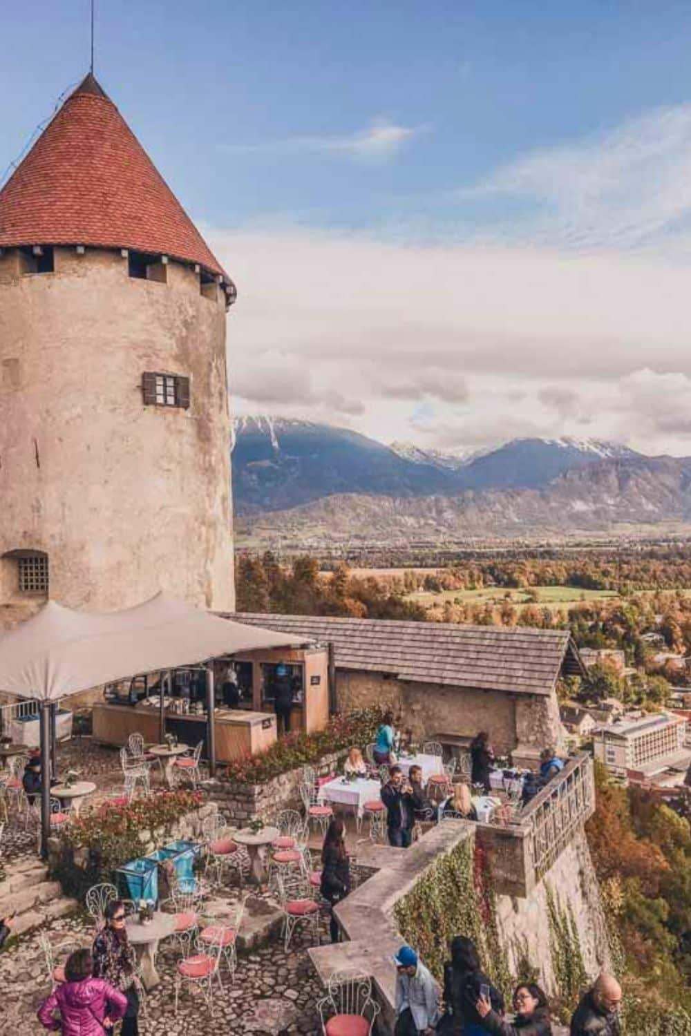 An outdoor terrace café located at the top of a hill, beside the tall stone tower of a castle, offers panoramic views over a picturesque landscape. Guests are seated at various tables, enjoying the scene under the shelter of a large white umbrella. Beyond the café, the expanse of Lake Bled is visible, surrounded by forested areas and mountains in the distance, under a vast sky with a mixture of clouds and blue patches.
