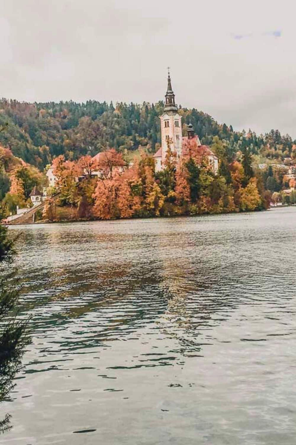 The picturesque Pilgrimage Church of the Assumption of Maria on Bled Island, surrounded by the autumnal hues of Lake Bled's trees, inviting visitors for a tranquil winter exploration.