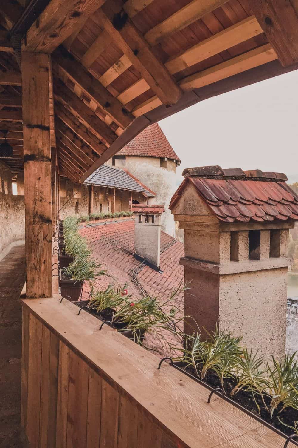 View from a wooden walkway within a castle, with wooden beams overhead and a traditional clay tile roof visible. Plants line the walkway's edge, and in the distance, a round castle tower looms against a pale sky. The architecture suggests a blend of functionality and ancient charm, typical of a medieval fortress.