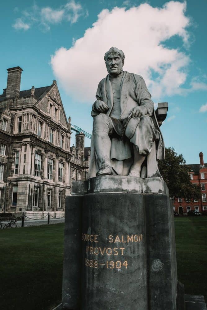 Bronze statue of George Salmon, Provost from 1888-1904, seated and holding a book, in front of the historic buildings of Trinity College Dublin under a clear blue sky with fluffy clouds.