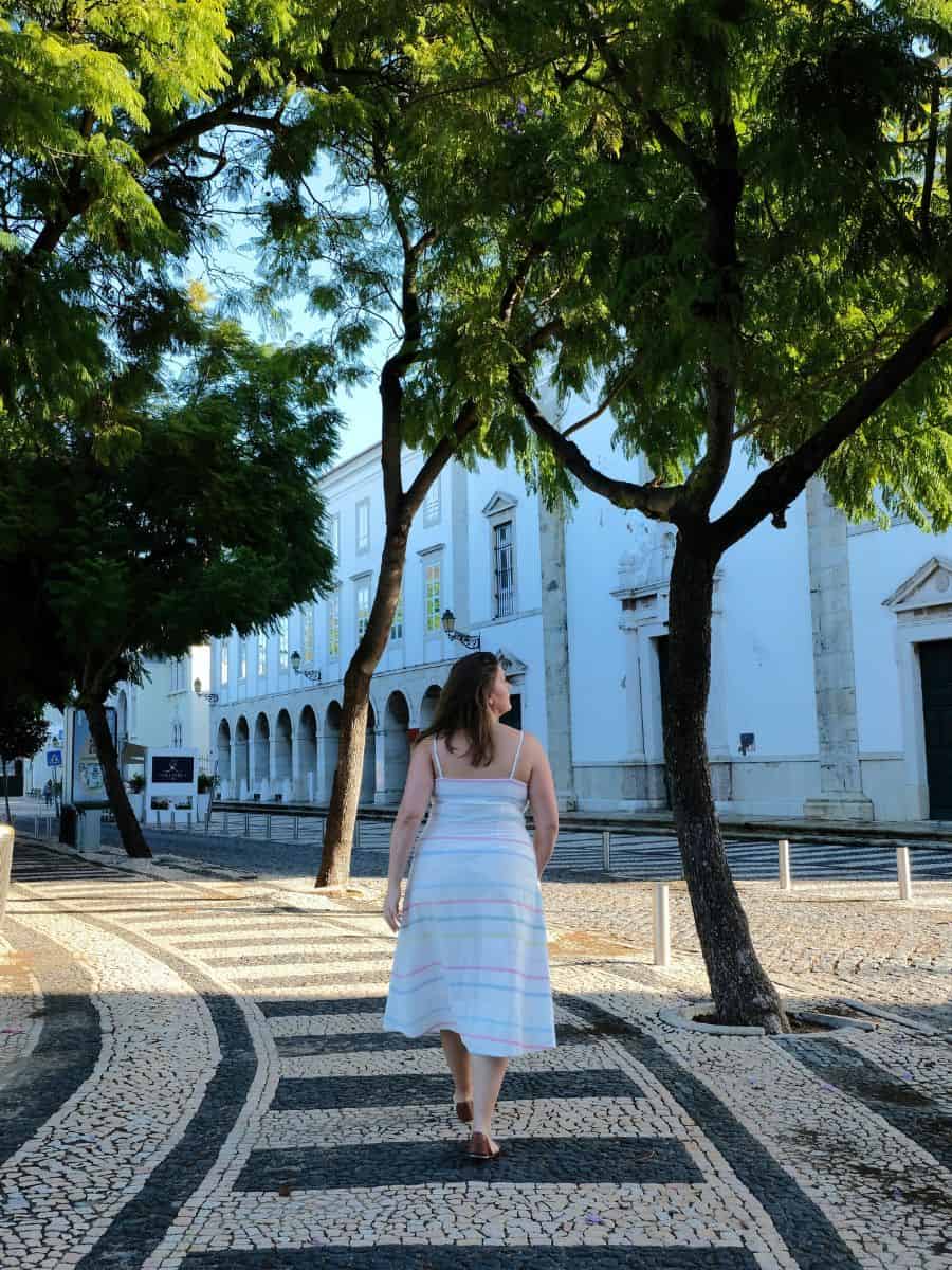 Woman enjoying solo travel in Faro, strolling along a picturesque cobblestone path lined with lush green trees, with traditional Portuguese white architecture in the background.