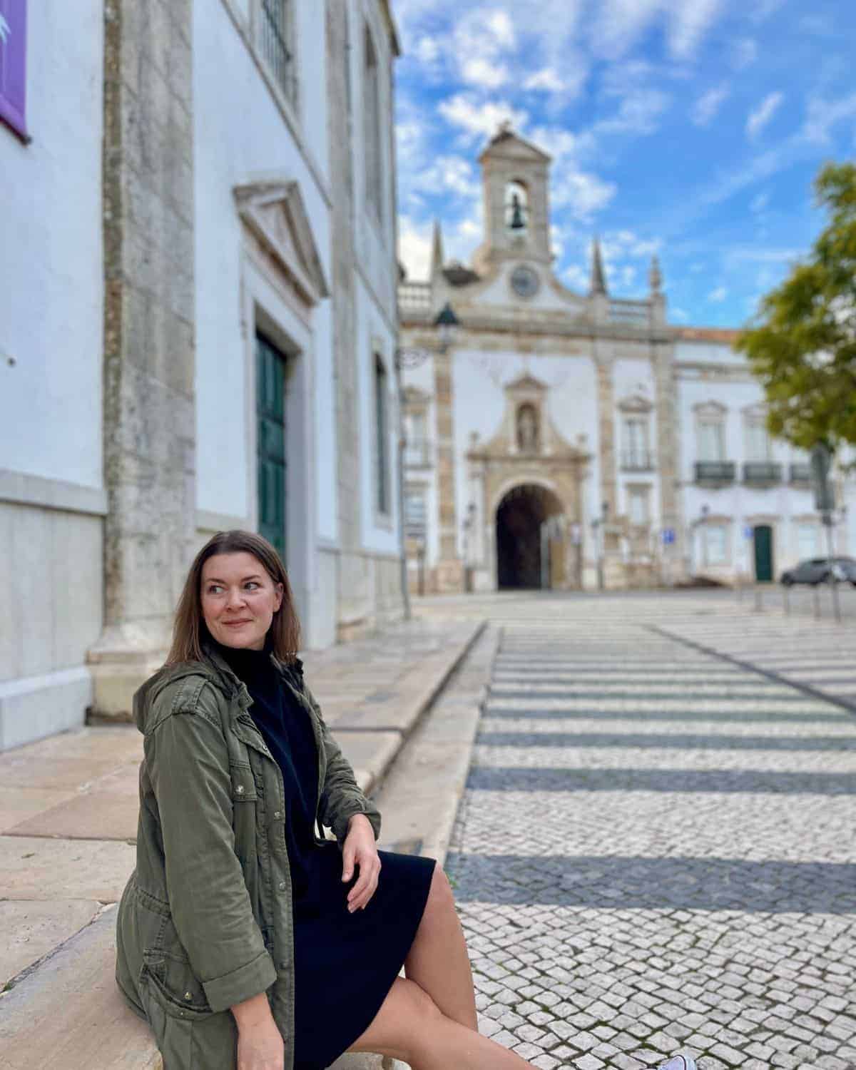 A woman sitting on the side walk with the old town of Faro in the background.