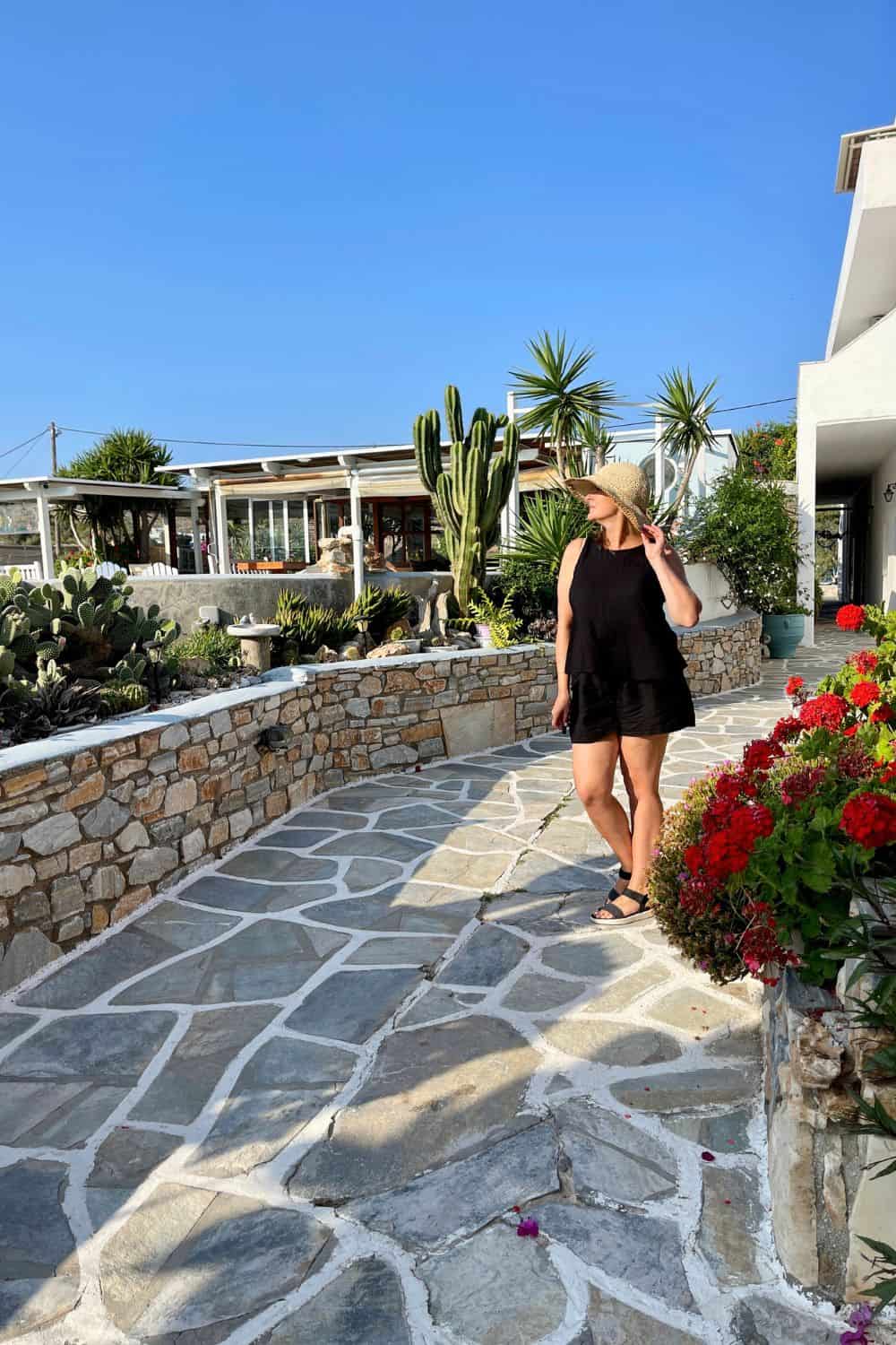 "A woman in casual summer attire and a straw hat strolls along a stone pathway surrounded by vibrant cacti and blooming flowers. In the background, a serene Greek island landscape with a white-washed building boasting a covered patio under a clear blue sky is visible, highlighting the region's unique architecture and flora.