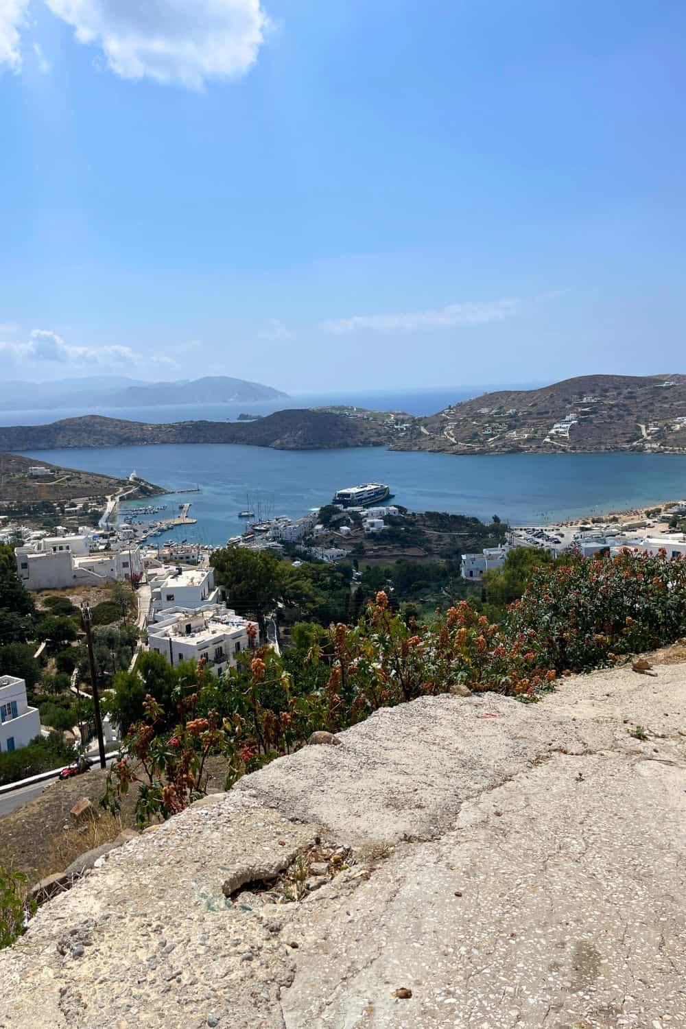 Panoramic view overlooking the harbor of Ios, Greece, with scattered white buildings nestled in the hilly landscape. The azure blue waters of the Aegean Sea create a calm bay, with boats moored in the marina and a ferry docked at the port. The foreground shows wild shrubs and plants typical of the Greek islands, thriving in the Mediterranean climate.