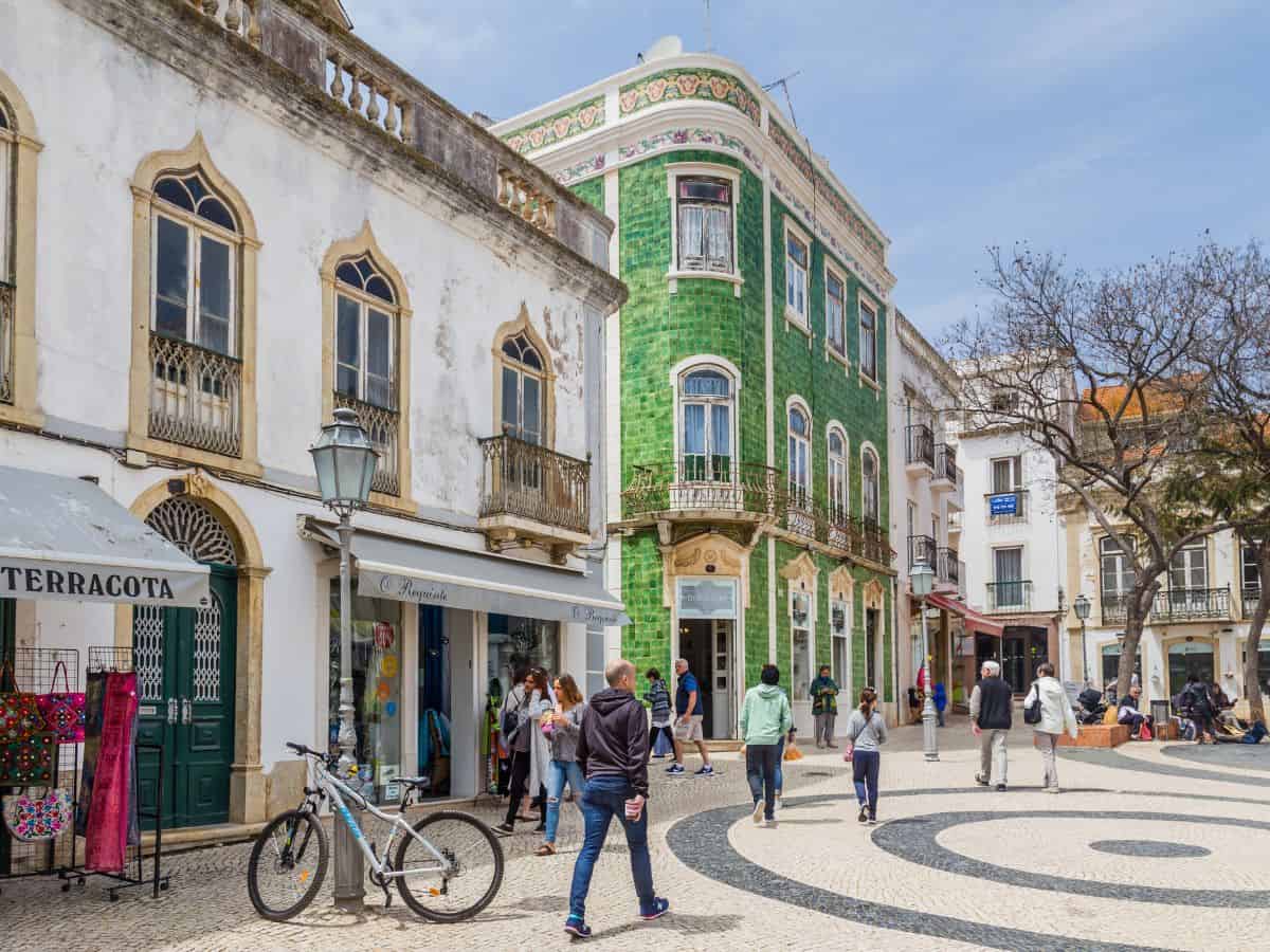 The vibrant town square in Lagos, Portugal, bustling with people and flanked by shops and the distinct green tiled façade of a traditional Portuguese building, encapsulating the lively atmosphere.