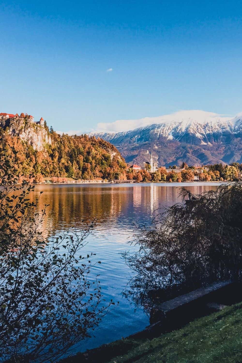 The picturesque Pilgrimage Church of the Assumption of Maria on Bled Island, surrounded by the autumnal hues of Lake Bled's trees, inviting visitors for a tranquil winter exploration
