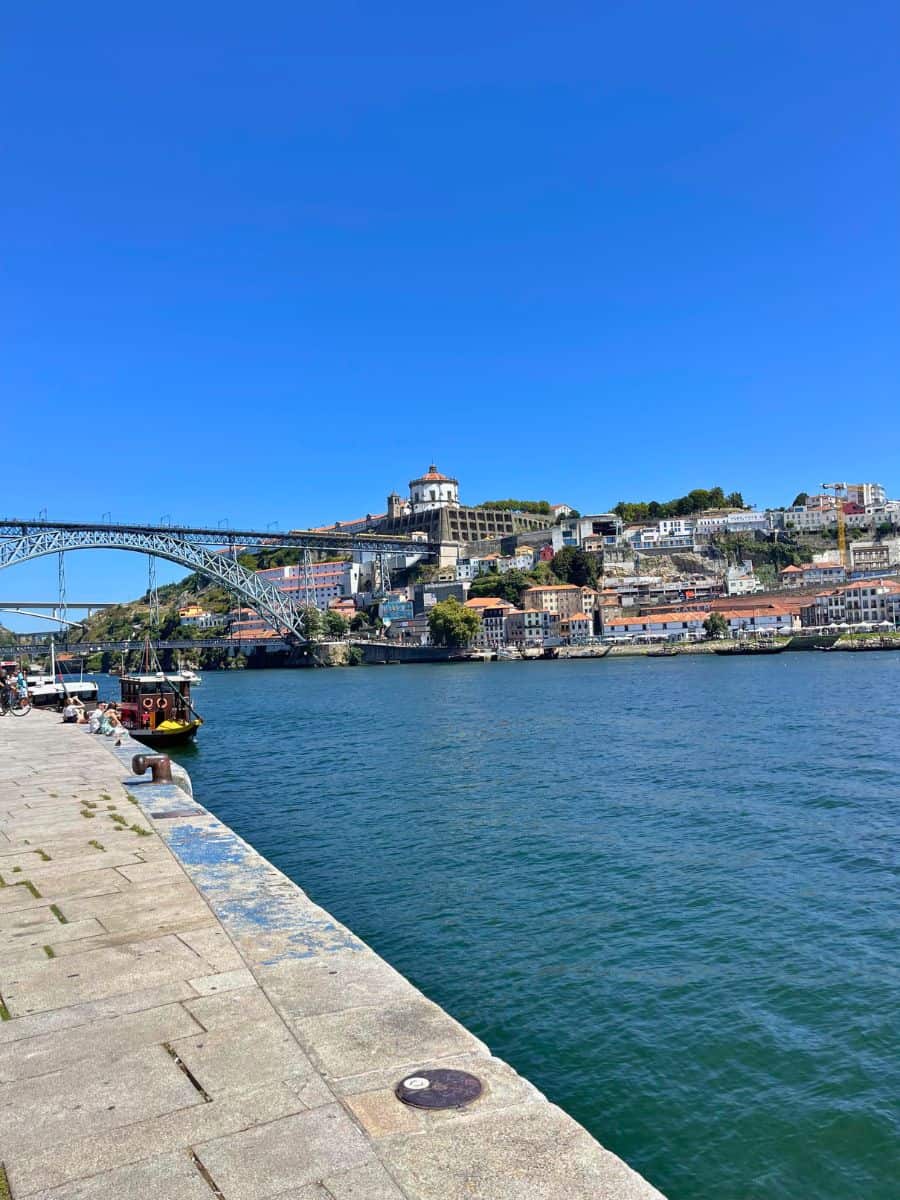 A breathtaking view of Porto's historic cityscape as seen from the riverbank, featuring the iconic Dom Luís I Bridge and the hilltop monastery, under a clear blue sky, a beautiful scene to witness on a day trip from Faro.