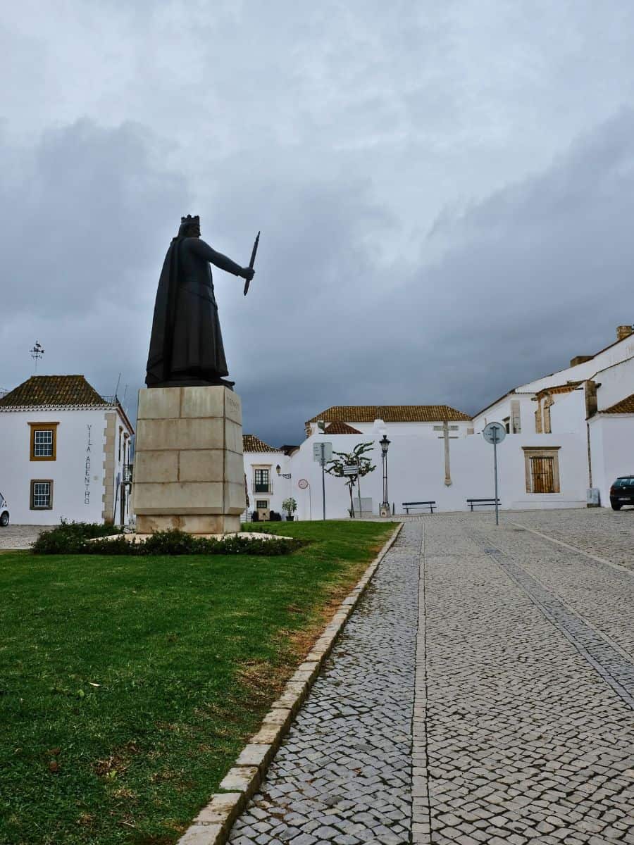 A towering statue of a historical figure brandishing a sword against a moody sky dominates the scene in Faro, with traditional white-washed buildings and cobblestone streets completing the historic atmosphere.