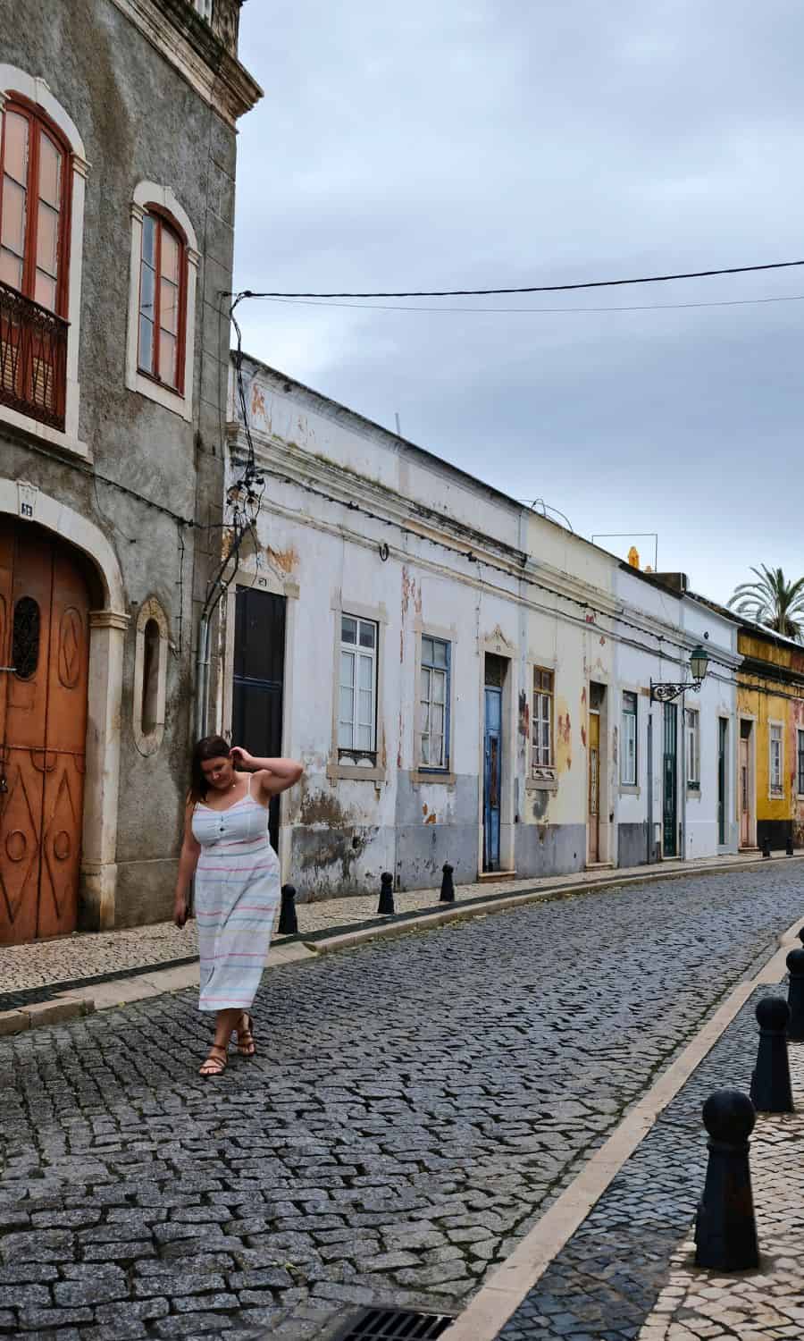 Woman solo traveler touching her hair while exploring the rustic charm of Faro's weathered buildings and cobbled streets, encapsulating the serene atmosphere of the city.