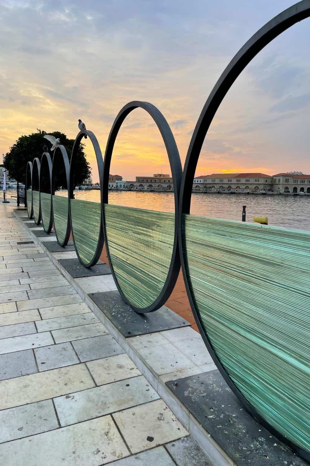 Sunset view at a waterfront in Syros with artistic urban design featuring a series of metal arches over a curved glass bench. The bench reflects the warm tones of the sky at dusk, while seagulls perch atop the sleek, modern structure. In the distance, the silhouette of neoclassical buildings lines the calm harbor, contributing to a peaceful and picturesque evening atmosphere.