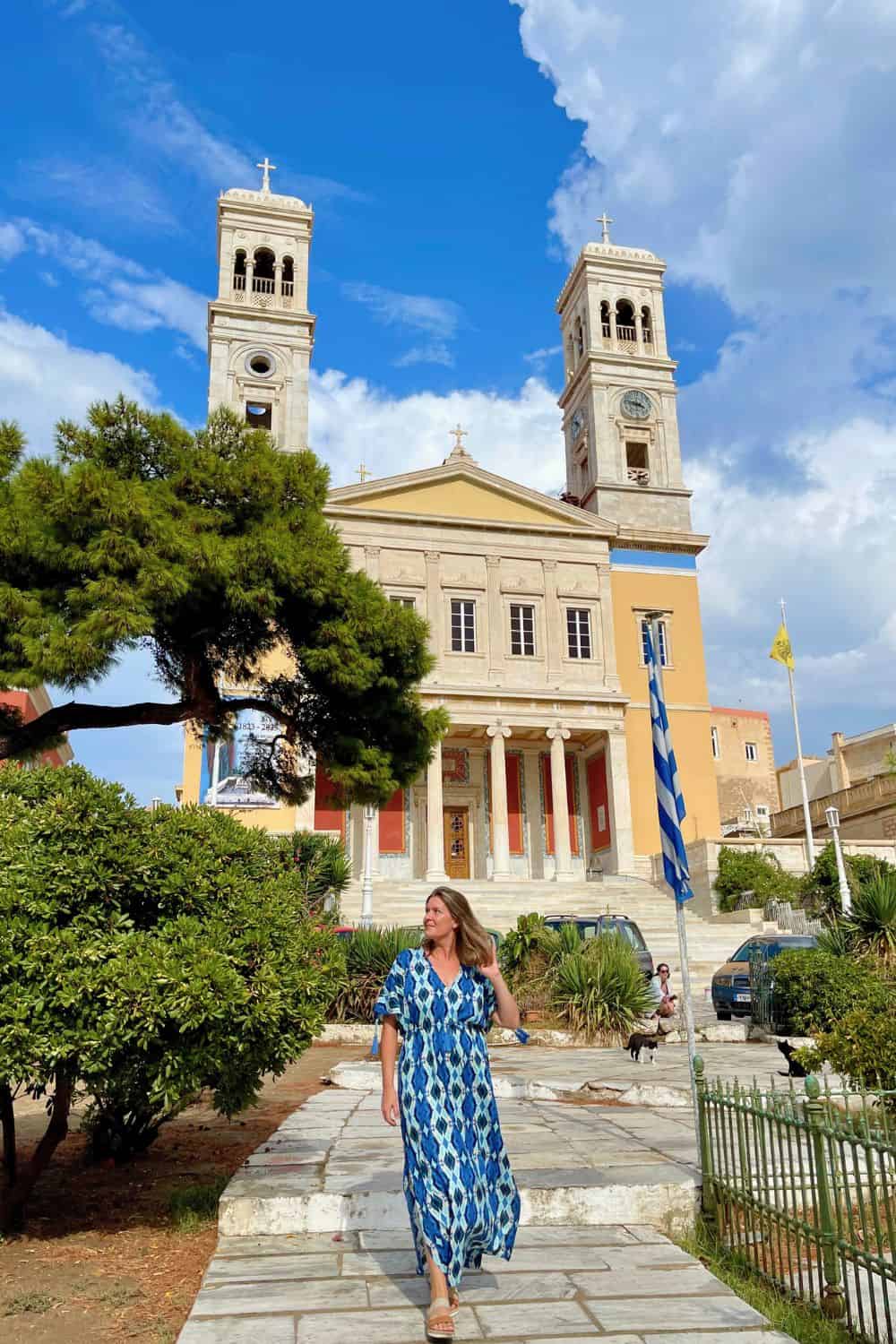 Large Greek Church in the background as a woman in a blue dress walks toward the camera. 