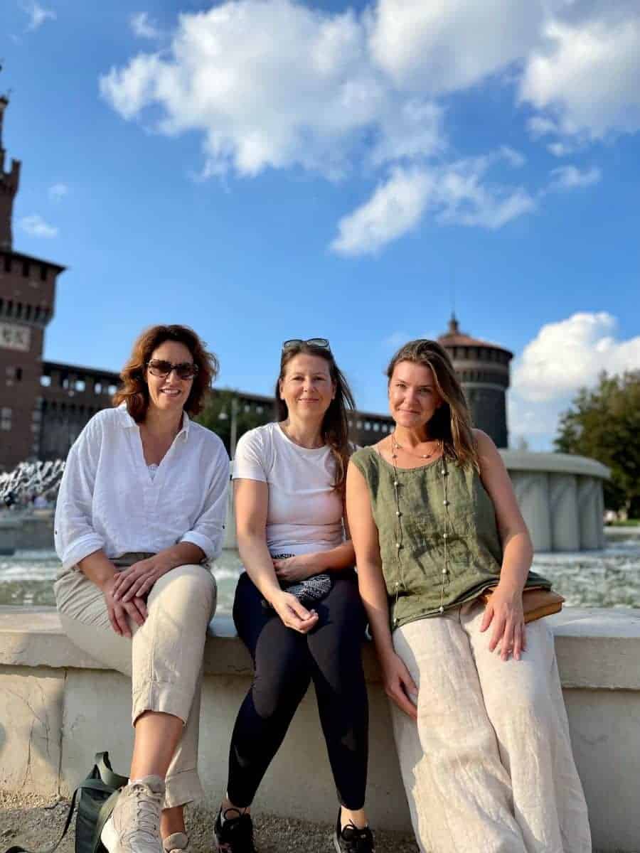 Three women seated side by side, enjoying a sunny day by the fountain at Sforza Castle in Milan, capturing the essence of friendship and travel.