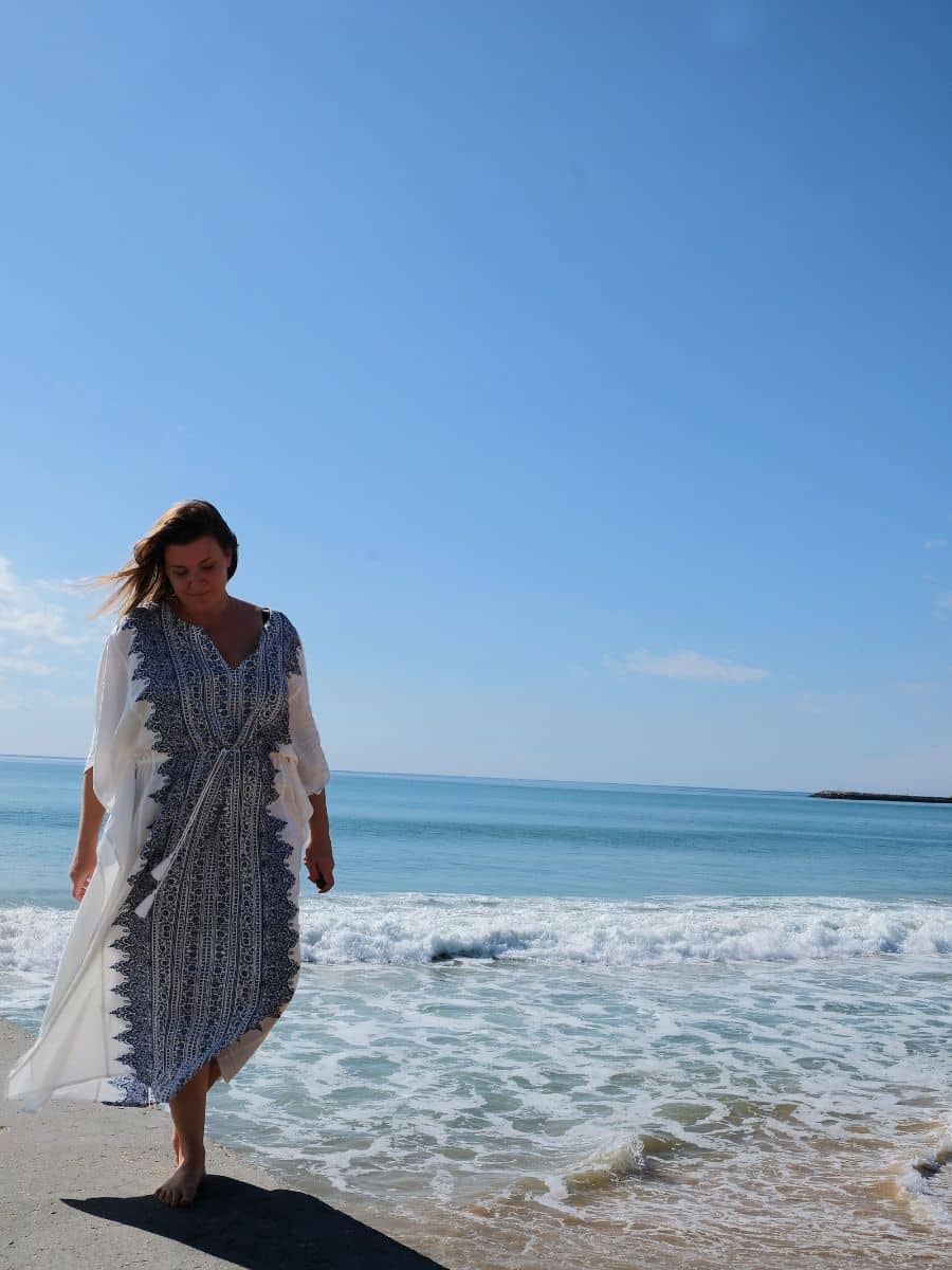 A woman walks along the shoreline, her white and blue embroidered dress flowing in the sea breeze, with the clear blue ocean stretching to the horizon, capturing the serene experience of a beach day trip from Faro.