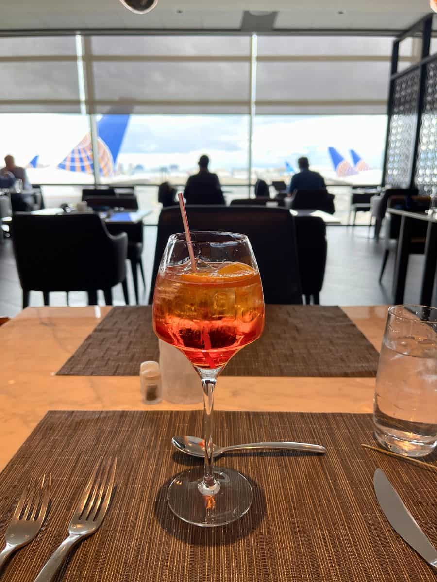 Foreground focus on a refreshing Aperol Spritz cocktail at an airport lounge dining table, with a serene view of airplane tails at the gates visible through the large window, encapsulating the exclusive travel experience offered by Priority Pass.