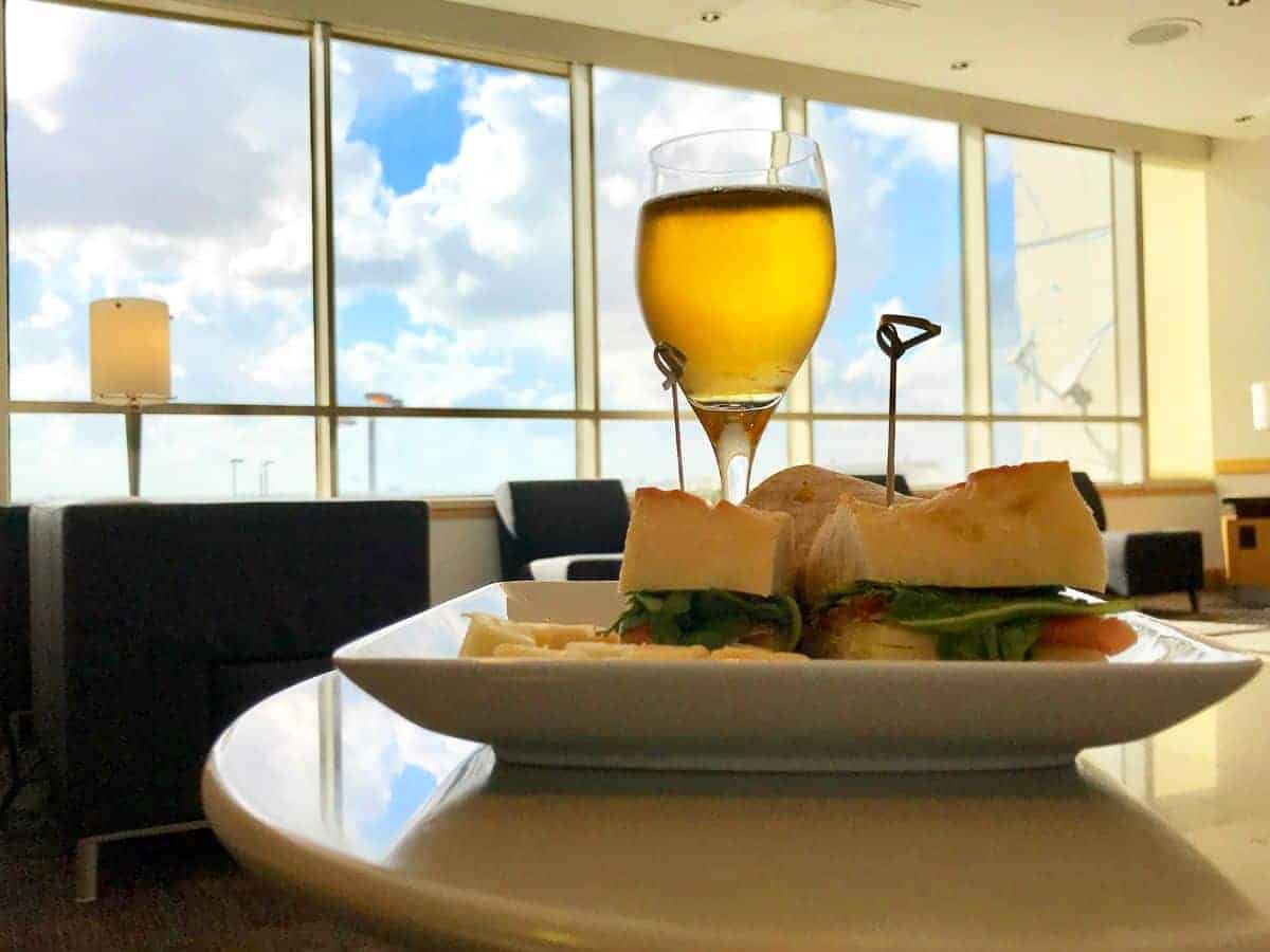A gourmet sandwich and a glass of amber beer set on a table in an airy airport lounge, with a panoramic view of the tarmac and a clear blue sky through floor-to-ceiling windows, reflecting the upscale experience offered by Priority Pass lounges.