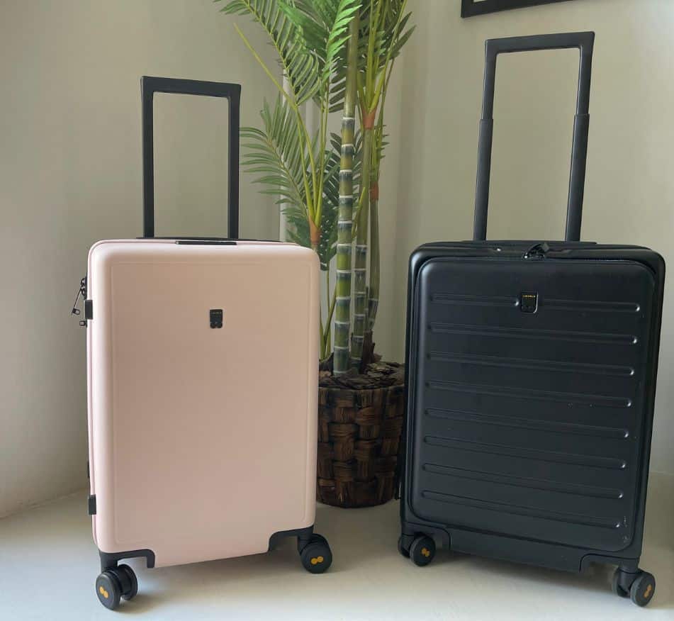 Two suitcases standing next to a potted palm tree indoors, one pale pink and one black, likely representing a promotion for Black Friday deals on luggage