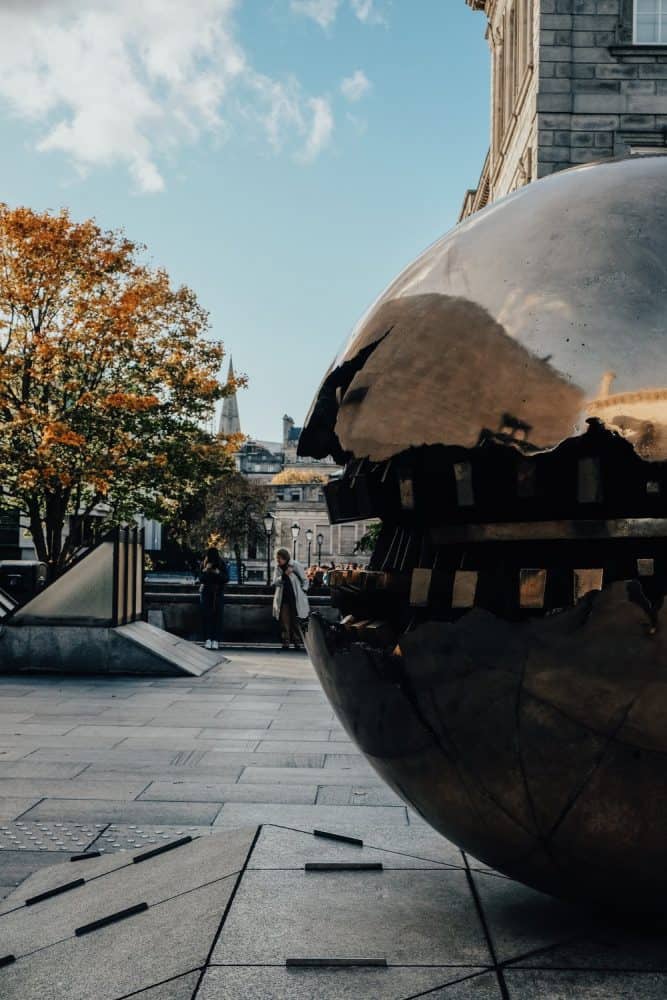 Close-up of the iconic 'Sphere Within Sphere' sculpture by Arnaldo Pomodoro at Trinity College Dublin, with a backdrop of autumn-colored trees and historic campus buildings under a bright blue sky.